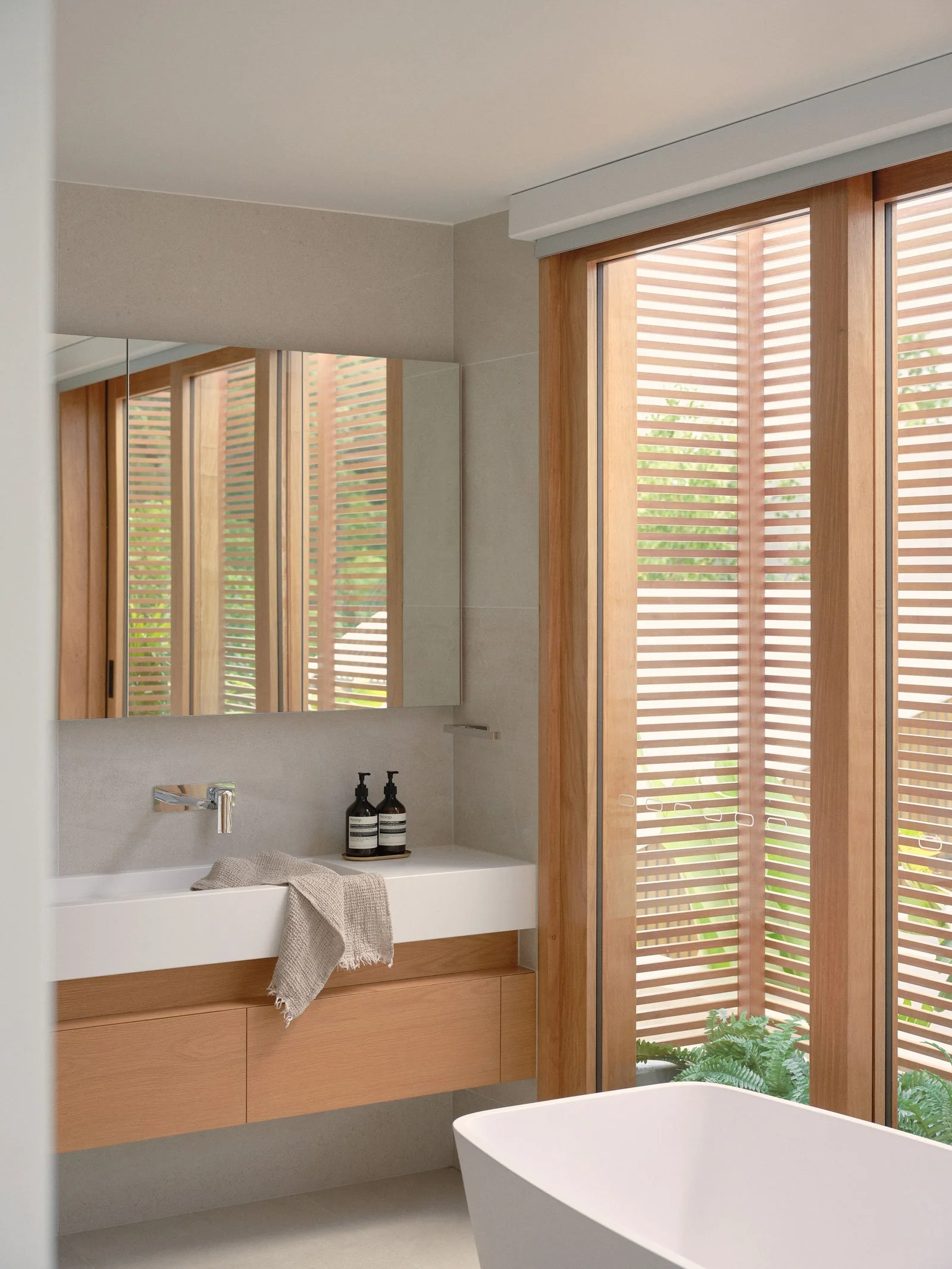 Modern bathroom with a floating wooden vanity, a rectangular white sink, black soap dispensers, a beige towel, a large mirror, and wooden window shutters with greenery outside.