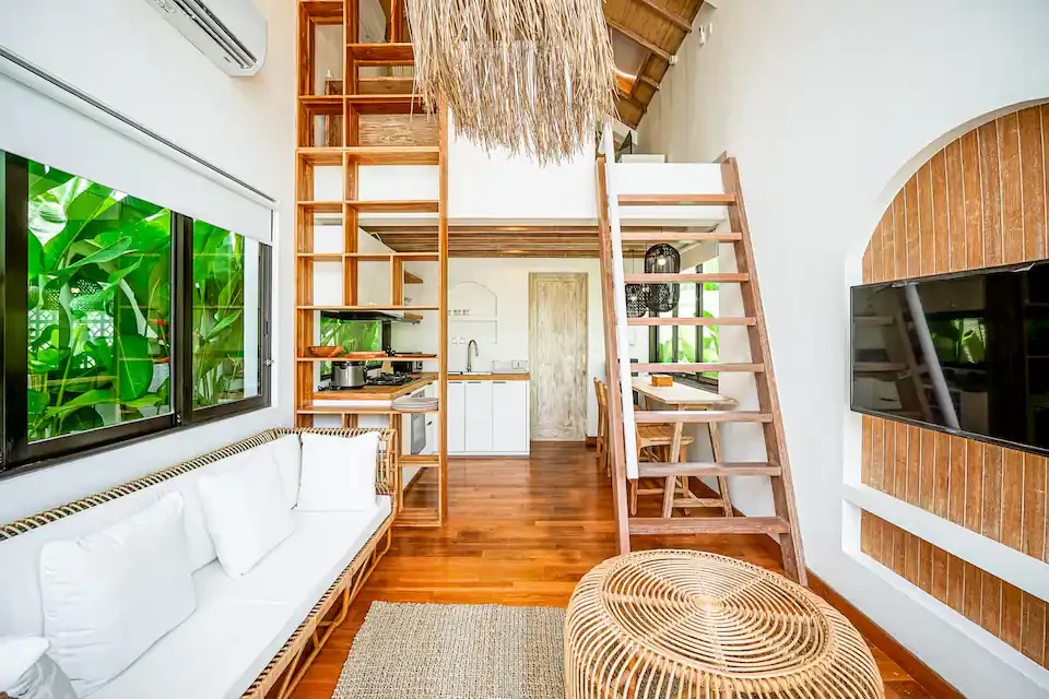 Living room with white sofa, wooden ladder leading to a loft, wooden dining table, flat-screen TV on a brick wall, windows showing green foliage, and wooden flooring.
