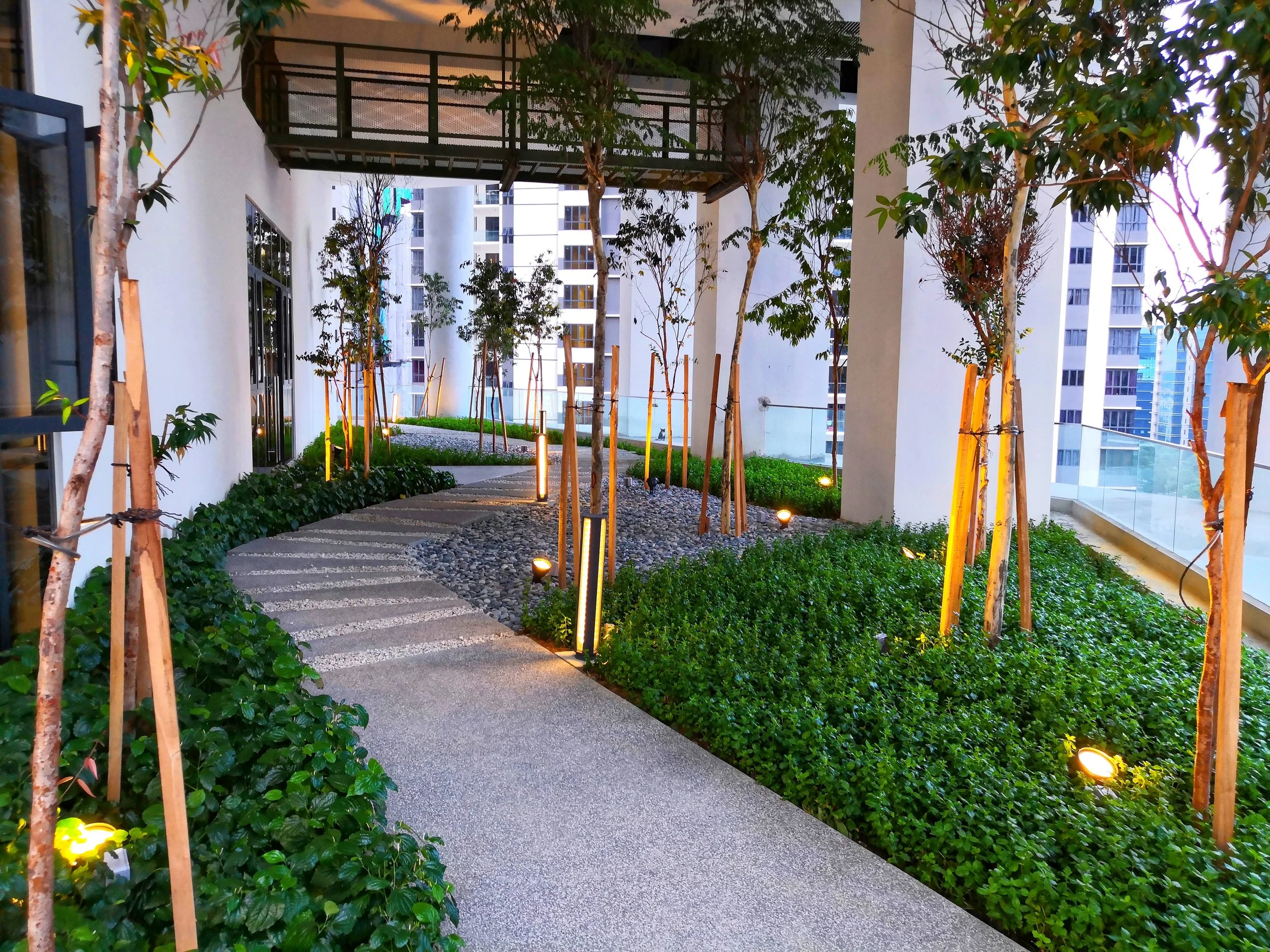 Urban balcony garden with a curved paved walkway, young trees supported by wooden stakes, green shrubs, and small lighting fixtures, in a high-rise residential building.