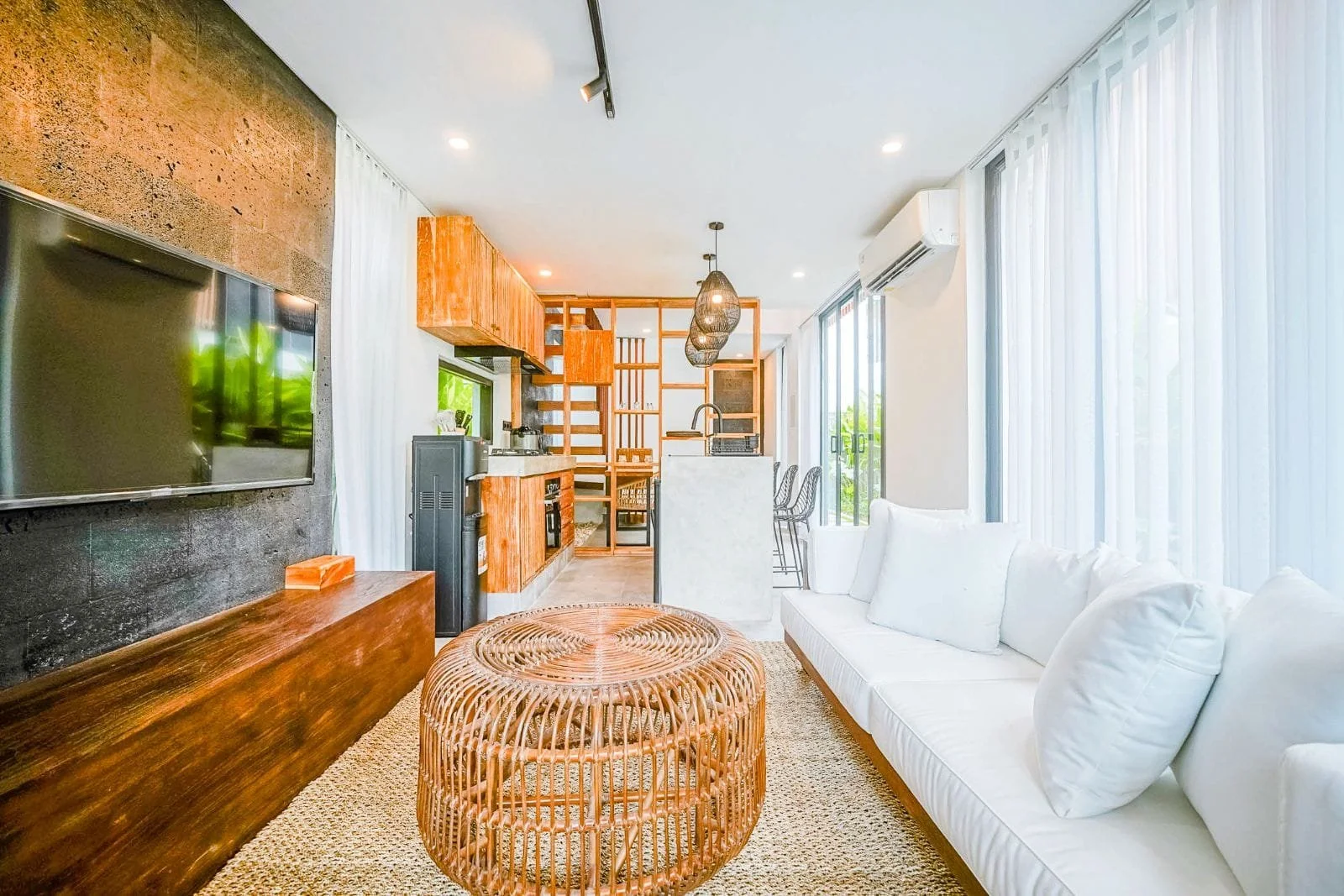 Bright living room with white sofa, natural woven coffee tables, wall-mounted TV, and large windows with white curtains. Kitchen area with wooden cabinets and bar stools visible in the background.