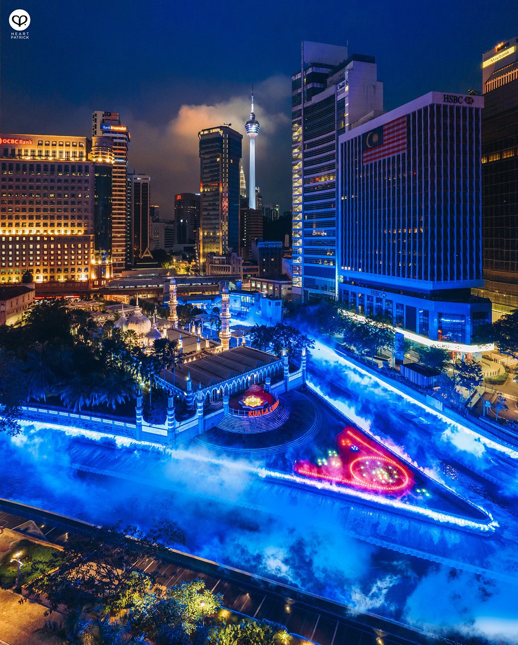 Nighttime cityscape of Kuala Lumpur, Malaysia, with illuminated skyscrapers, the KL Tower, and a misty, colorful fountain display in the foreground.