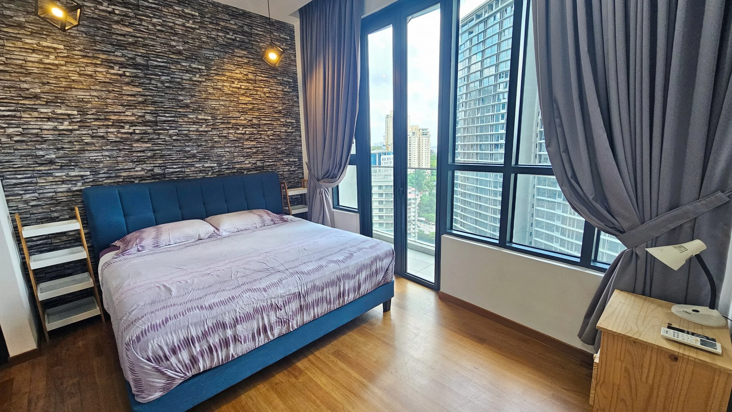 Modern bedroom with a large bed having a blue upholstered headboard, purple striped bedding, and two pillows. The room features a stone accent wall, wooden floors, and large windows with gray curtains, showing a cityscape with tall buildings and gree