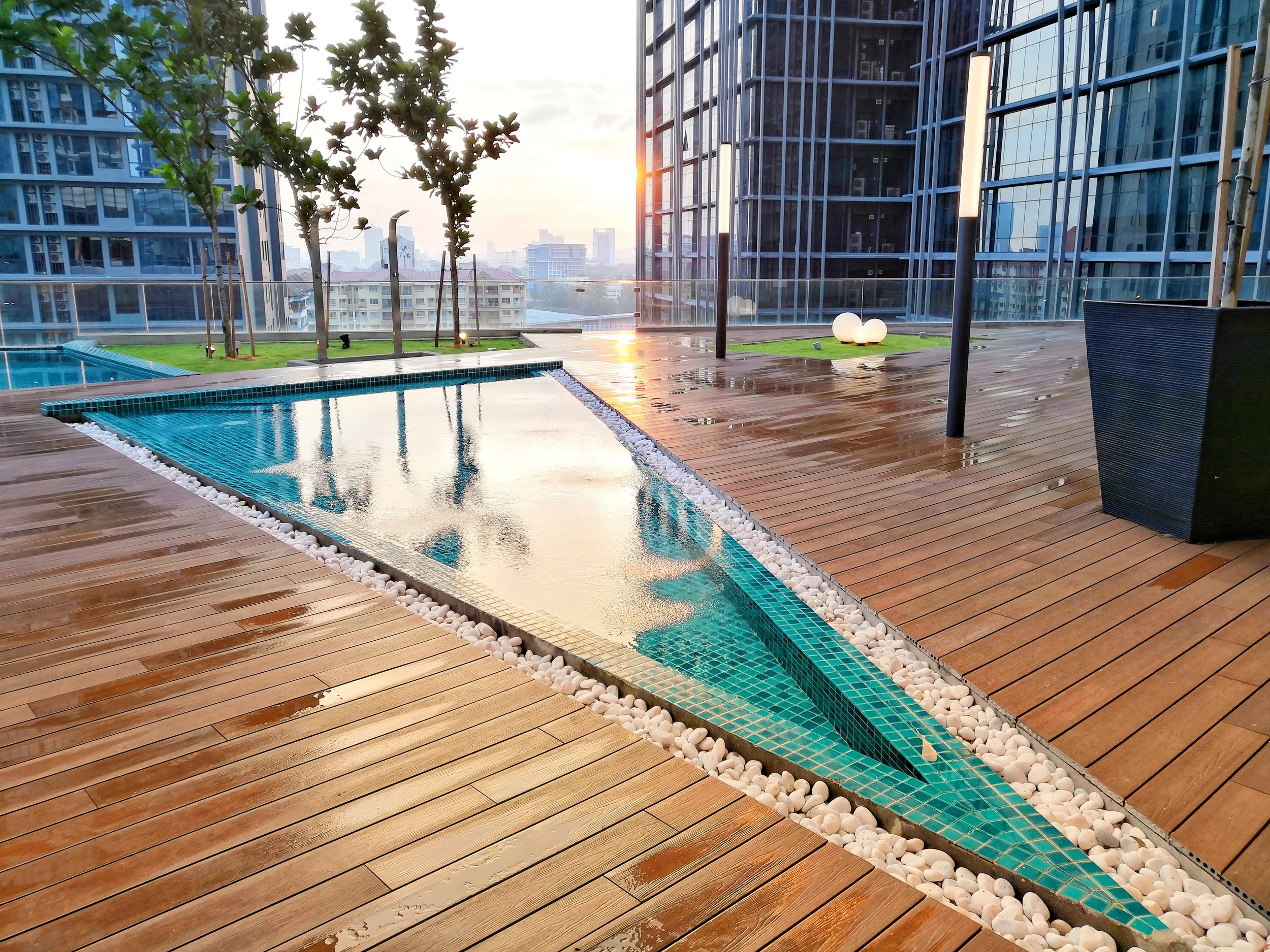 Modern rooftop pool with wooden deck, potted plants, and city skyline at sunset.