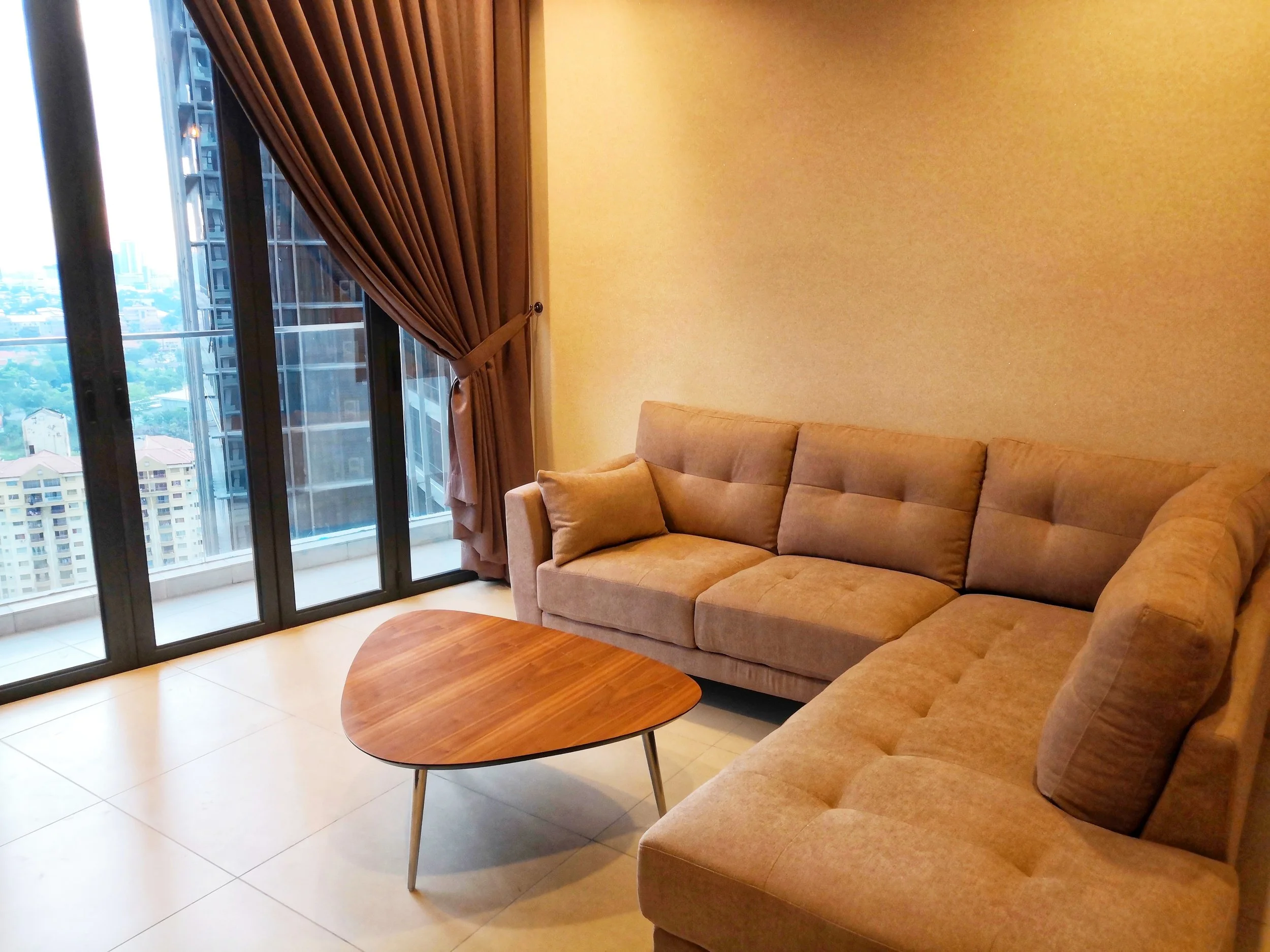 Living room with a beige sofa, a small wooden coffee table, a large window with a brown curtain, and a city view outside.