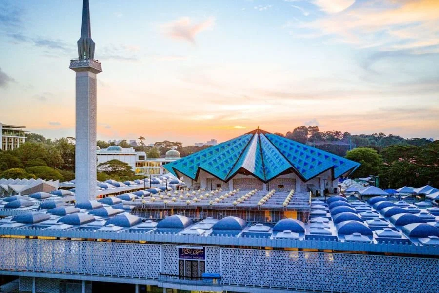 A mosque with a tall minaret and a blue, pyramid-shaped roof, surrounded by a patterned fence and likely in a city or town at sunset.