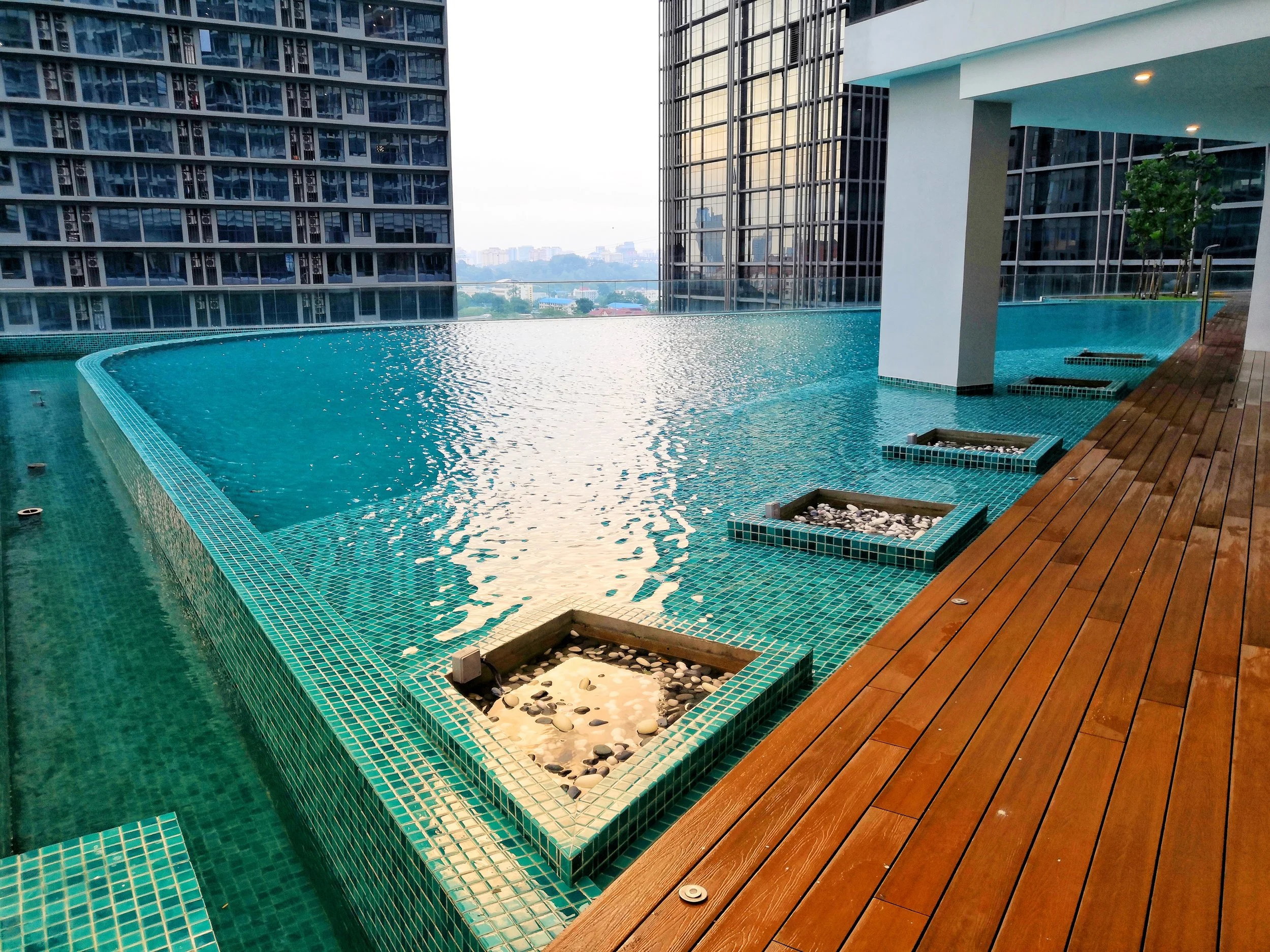 Rooftop infinity pool with wooden deck and city buildings in the background.