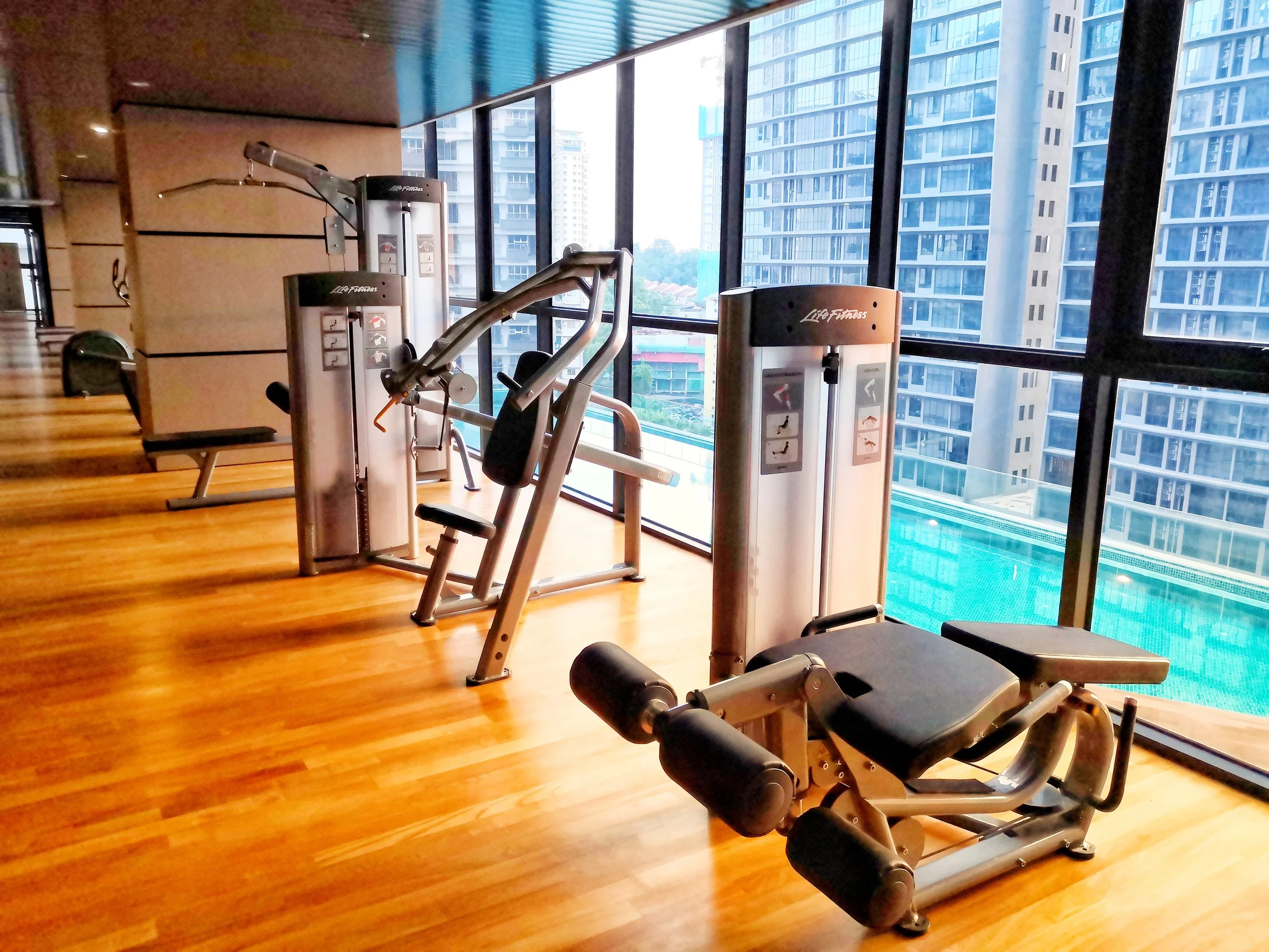 Indoor gym with exercise machines on wooden floor, large windows showing city buildings outside.