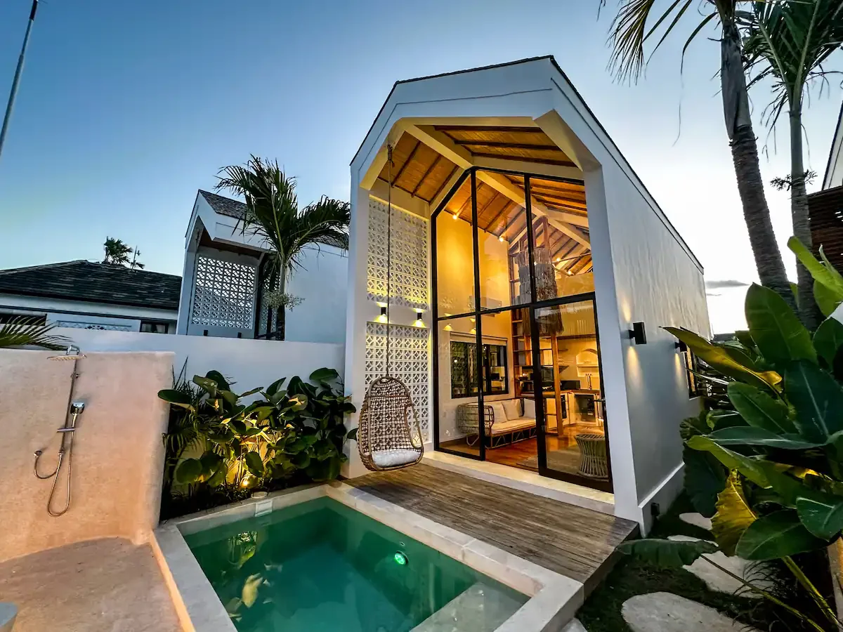 A modern tiny house with large glass doors, surrounded by tropical plants, next to a small swimming pool at dusk.