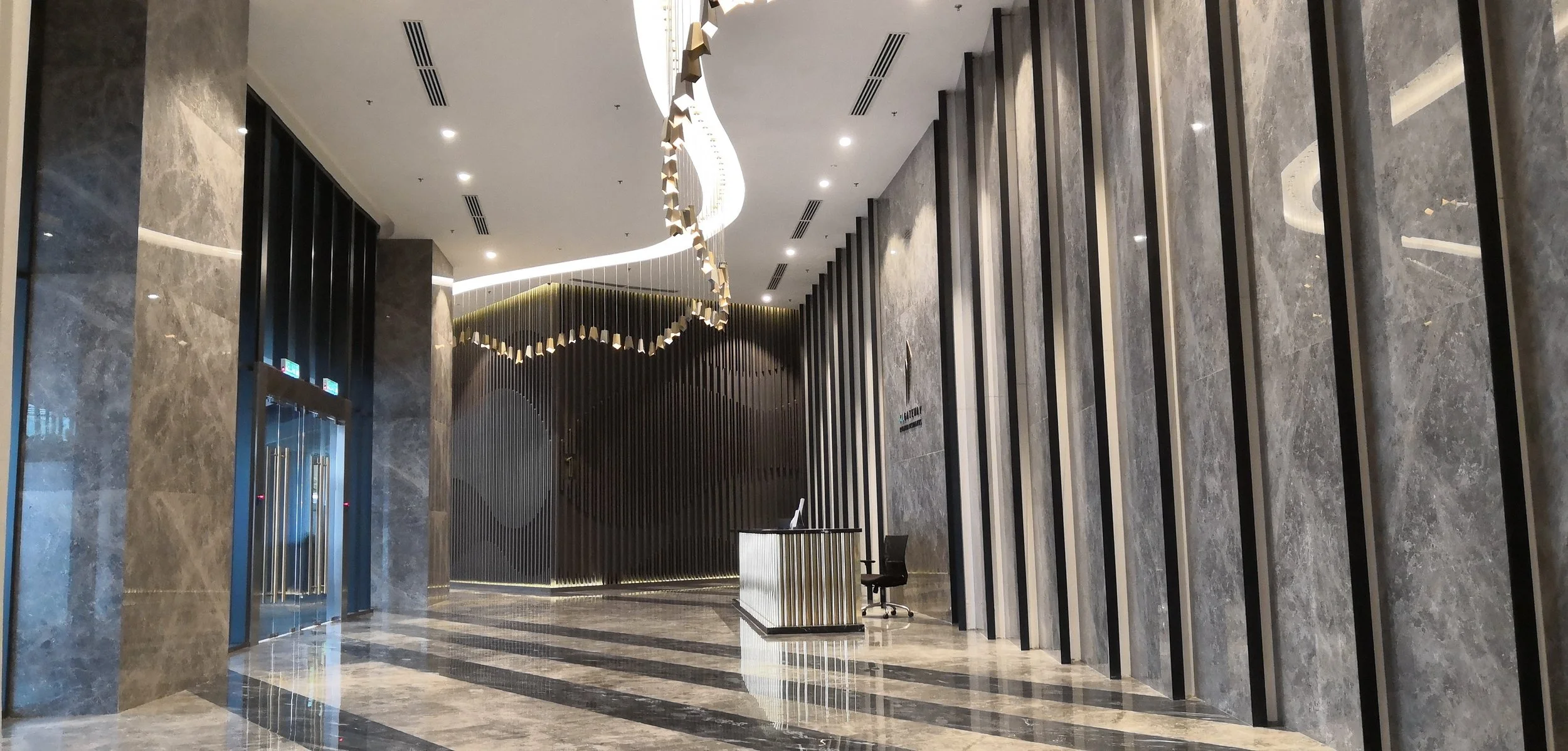 Modern hotel lobby with gray marble floors and walls, a reception desk, and decorative vertical black panels on the right wall. An artistic chandelier hangs from the ceiling, and there are large windows near the elevator doors on the left.