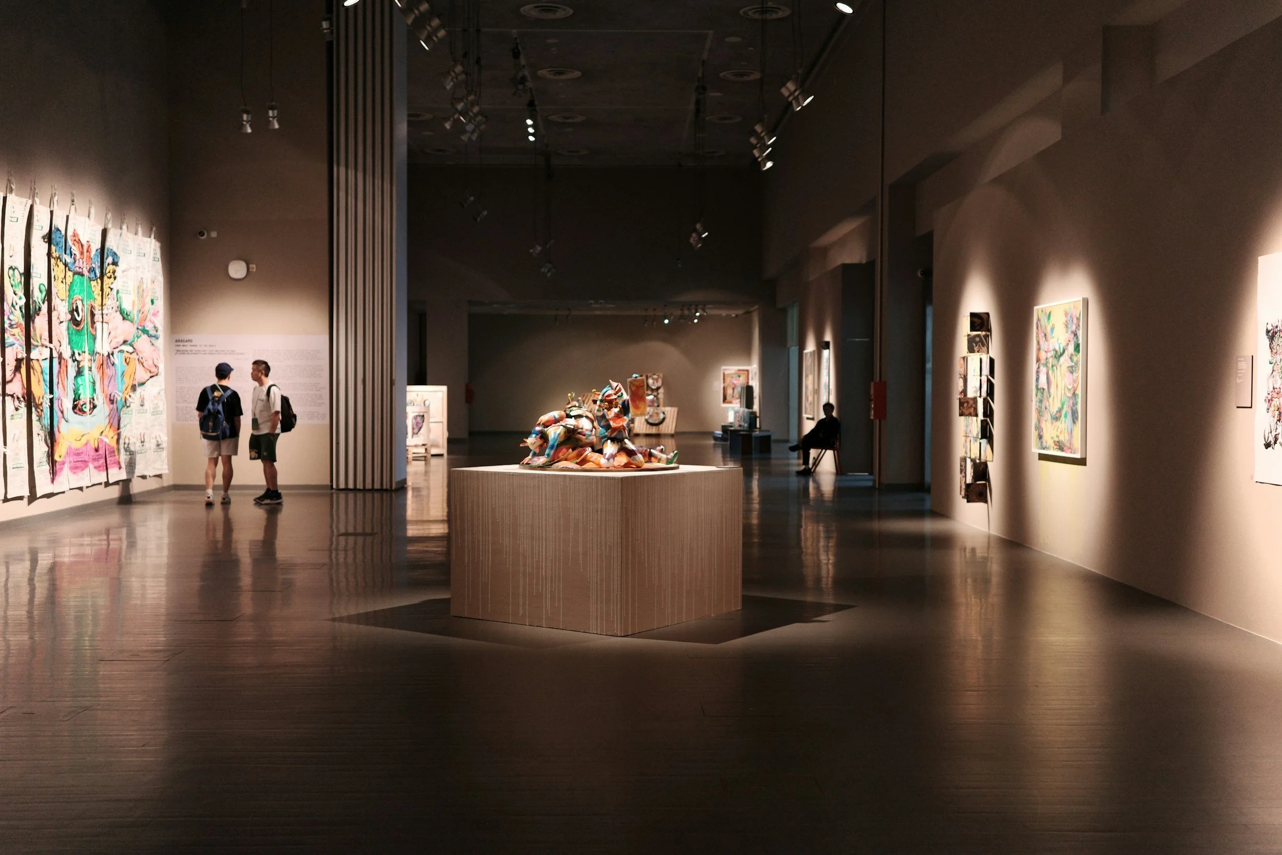 Interior of an art gallery with artworks displayed on walls and a sculpture on a central pedestal, with visitors viewing the exhibits.