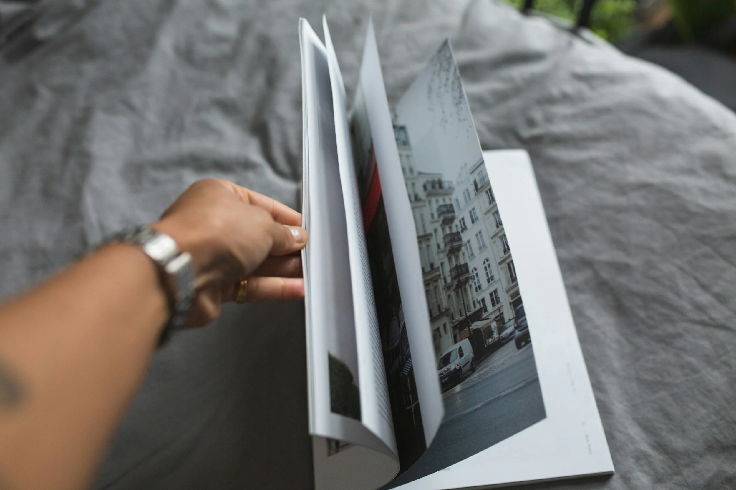 Open magazine featuring photos of buildings and city streets, held by a person's hand with a silver watch on wrist, placed on a soft gray fabric surface.