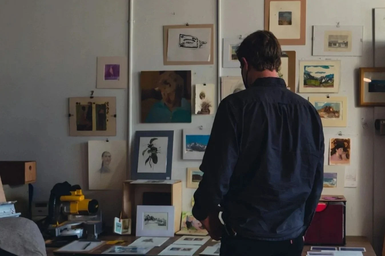 A man with dark hair, wearing a dark shirt, stands in an art gallery or studio, looking at various artworks displayed on the wall and table.