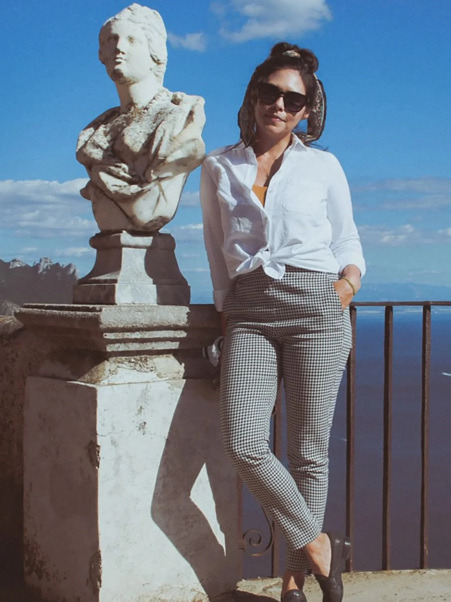 A woman with dark hair in a bun, wearing sunglasses, a white knotted blouse, checkered pants, and black shoes, leaning against a black metal railing beside a white stone bust sculpture of a woman with curly hair against a blue sky with some clouds.