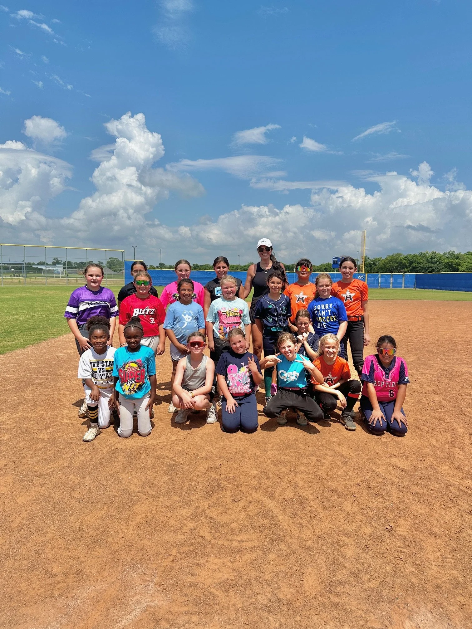Group of young girls after a clinic with The Softball Doctor, Jessica Bowden on a softball field under a partly cloudy sky.