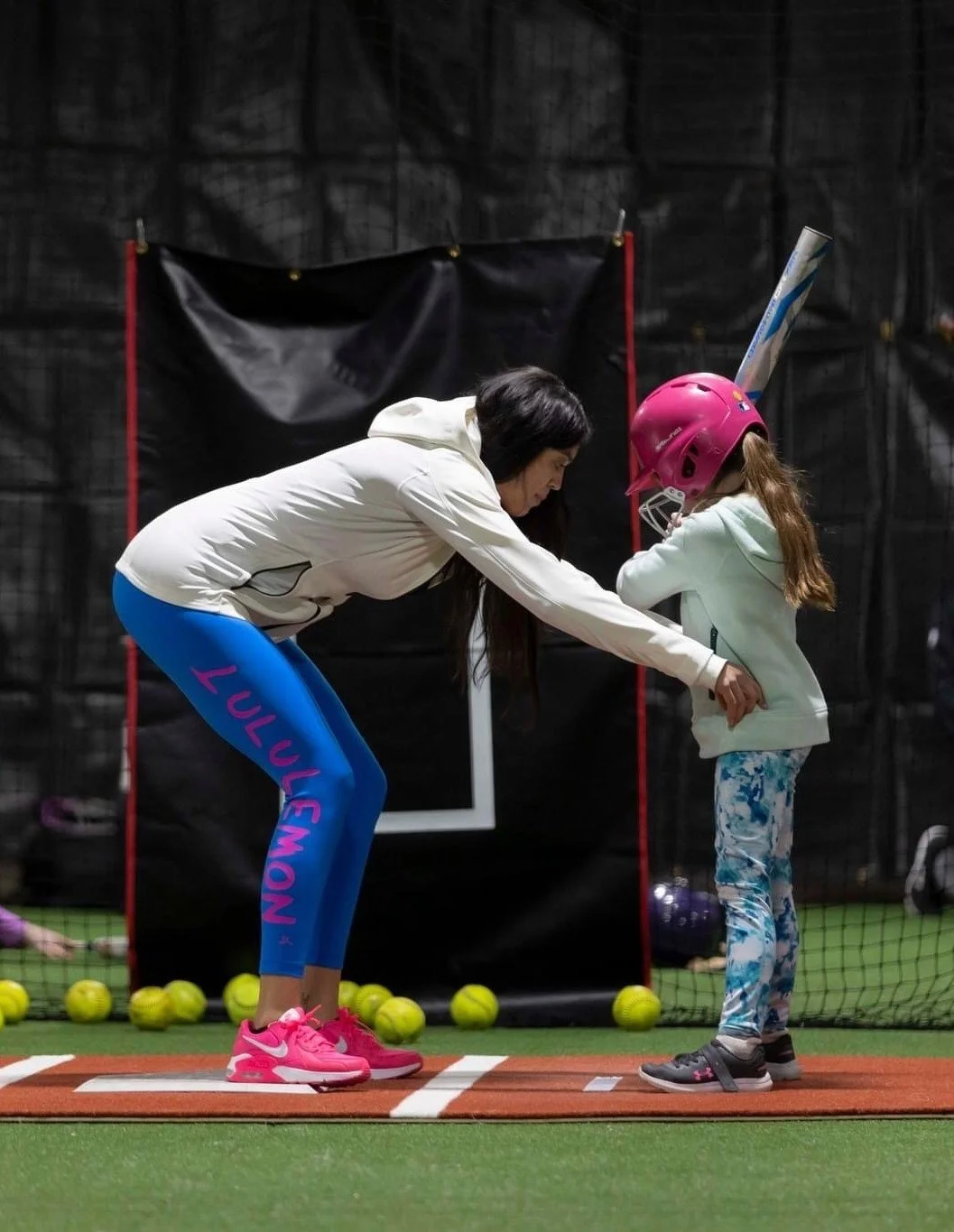 The Softball Doctor and a young girl are practicing baseball indoors. The Softball Doctor is showing the girl how to hold a baseball bat. The girl, wearing a pink helmet, is holding a bat.