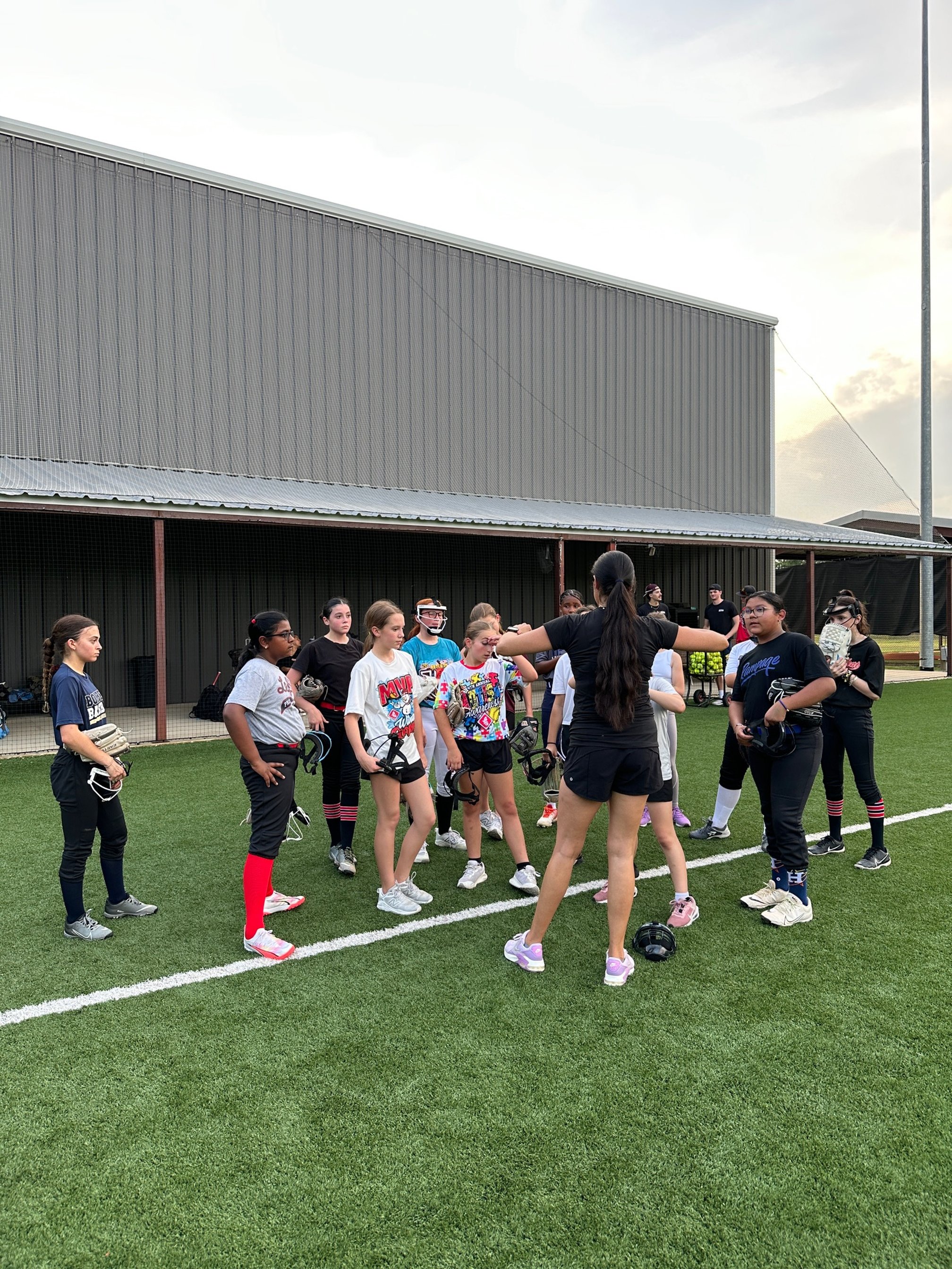 A group of young female softball players listening to The Softball Doctor, Jessica Bowden, on a field with artificial turf, holding their helmets and standing near the dugout.