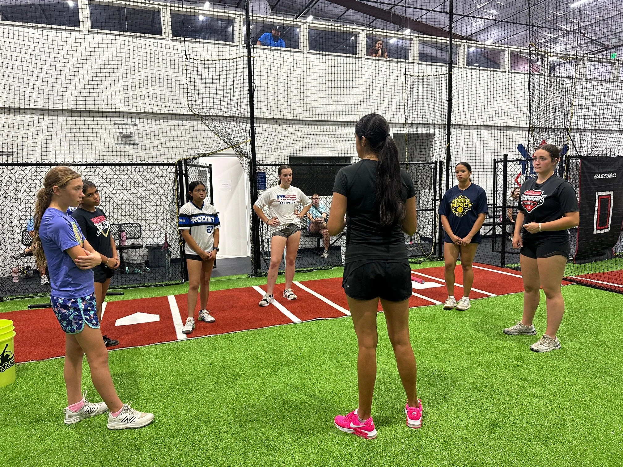 Group of young female athletes standing in a training facility, listening to The Softball Doctor, Jessica Bowden, at a hitting clinic.