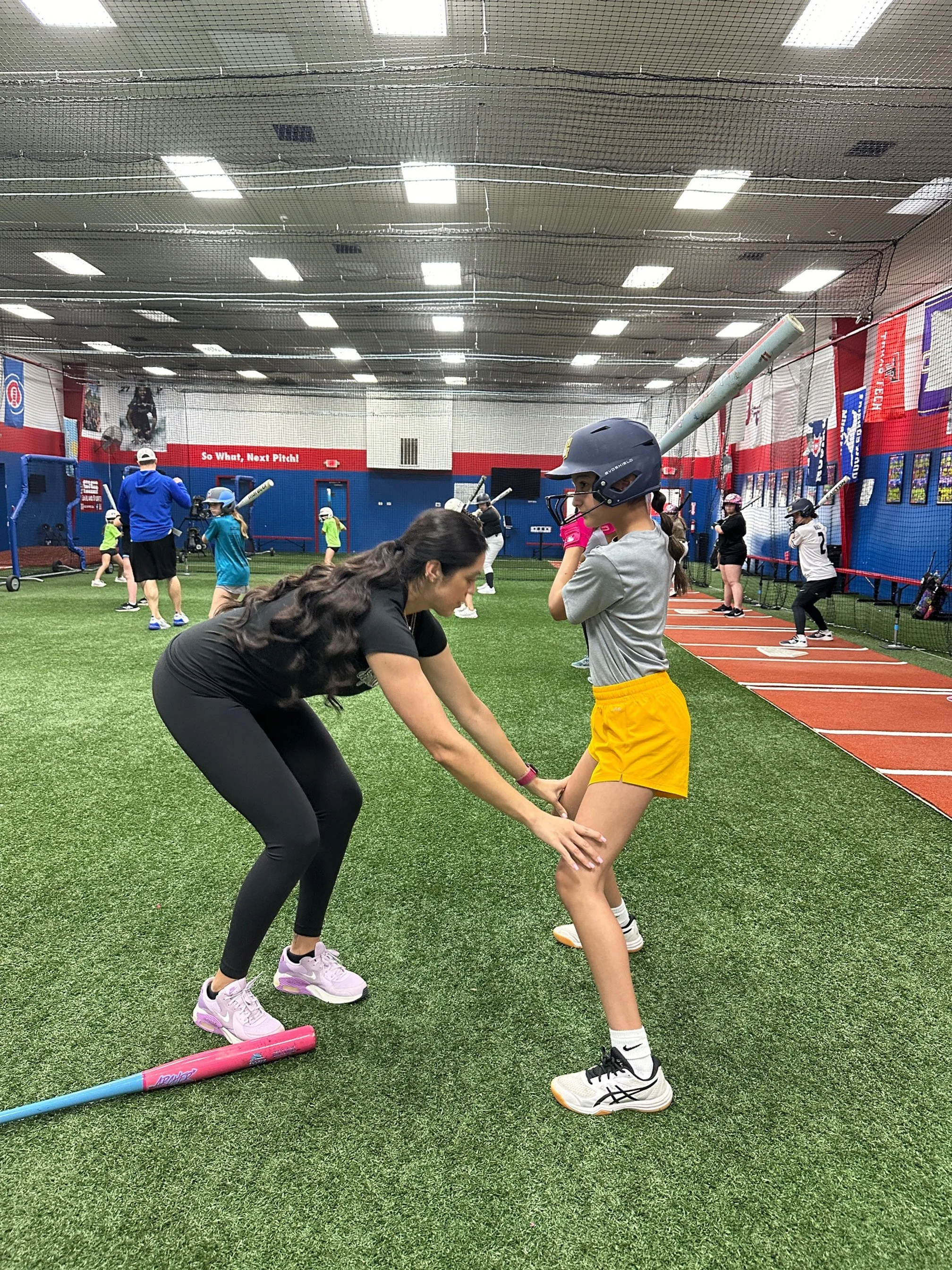 The Softball Doctor, Jessica Bowden, helping a young girl practice baseball batting at an indoor sports facility with artificial turf.