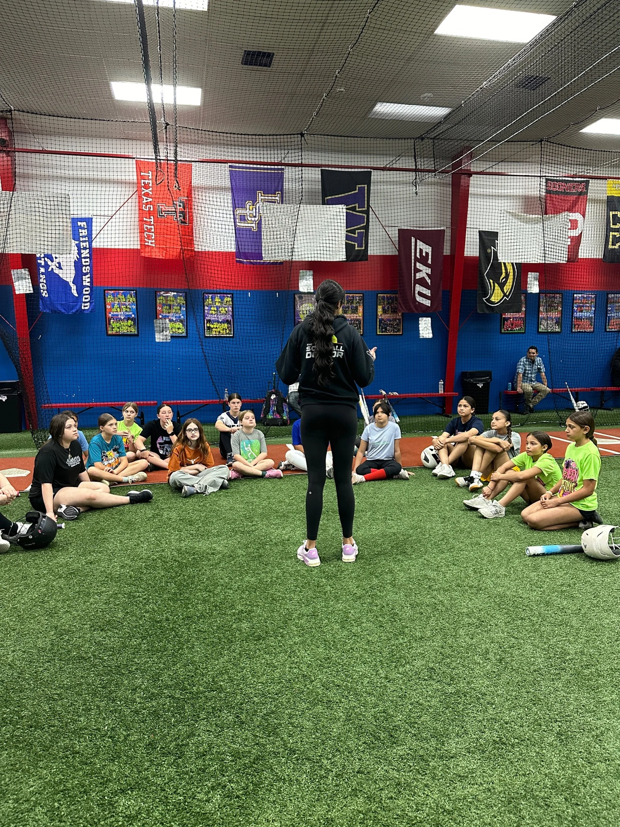 The Softball Doctor, Jessica Bowden, giving instructions to a group of young female athletes sitting on the artificial turf inside a sports facility. Flags and banners are hanging from the ceiling in the background.