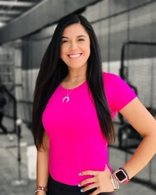 The Softball Doctor, Jessica Bowden, smiling, wearing a bright pink shirt, a necklace with a crescent moon, a smartwatch, and bracelets, standing outdoors.
