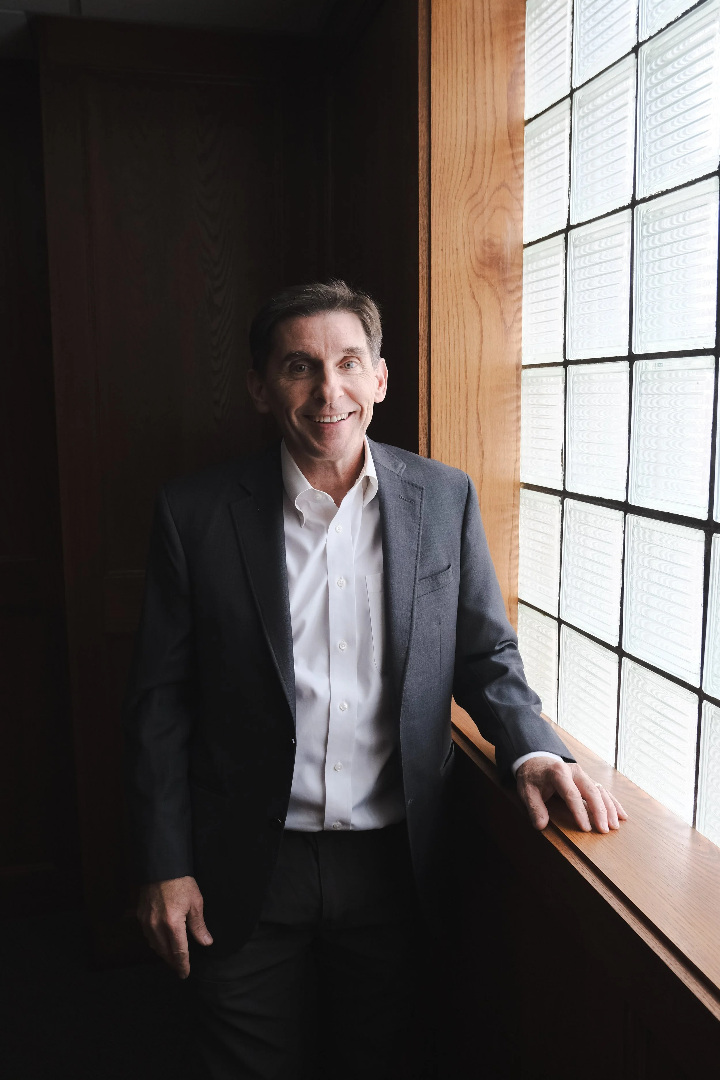A man in a business suit smiling, standing next to a window made of glass blocks, with wood paneling behind him.