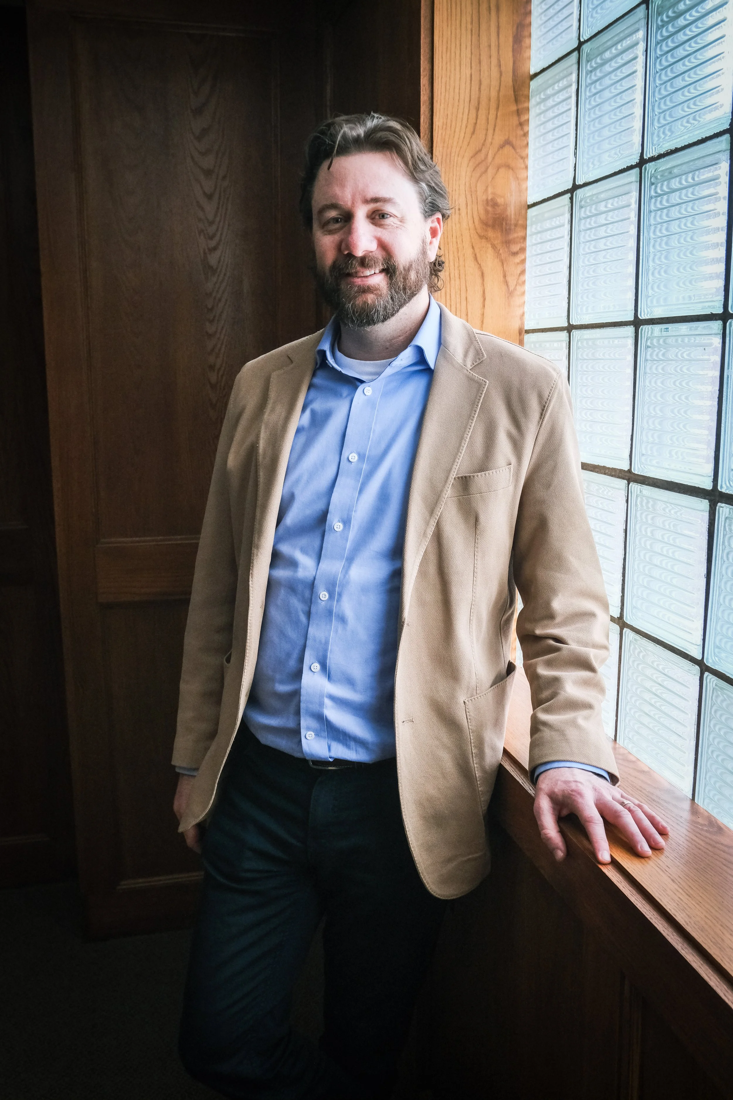 A man with a beard in a tan blazer and blue shirt standing indoors near a window with glass blocks.