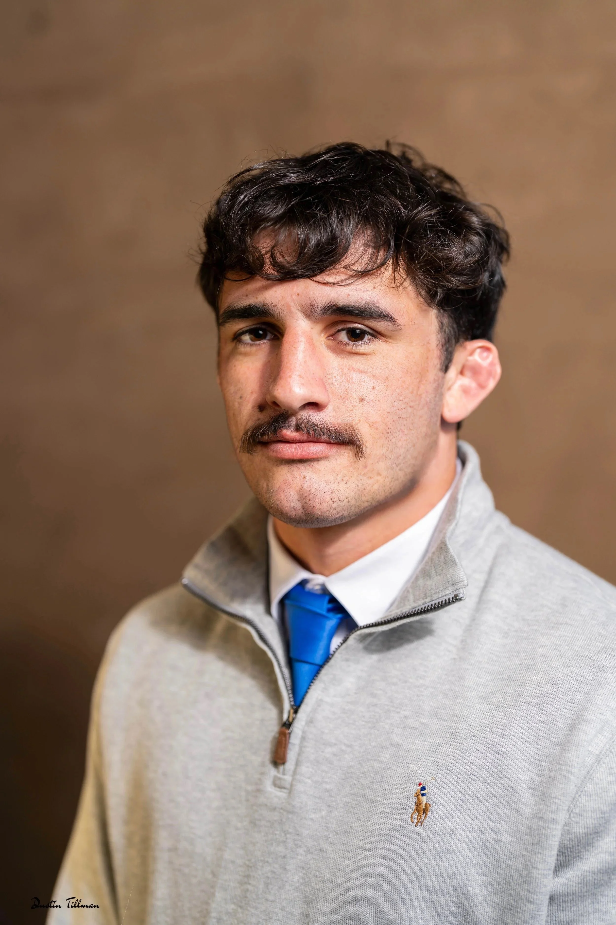A young man with dark, curly hair and a mustache, wearing a light gray quarter-zip sweater with a small Polo Ralph Lauren logo on the chest, a white dress shirt, and a blue tie, standing against a brown background.