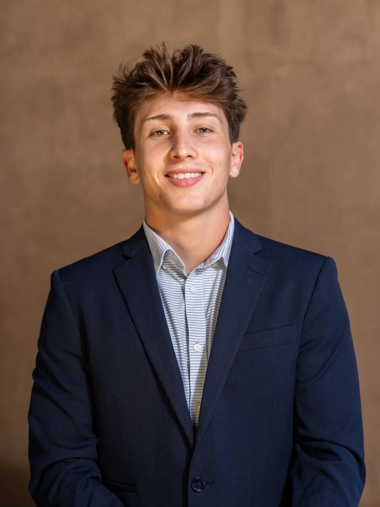Young man with brown, spiked hair wearing a navy blazer and light-colored striped shirt, smiling against a plain brown background.