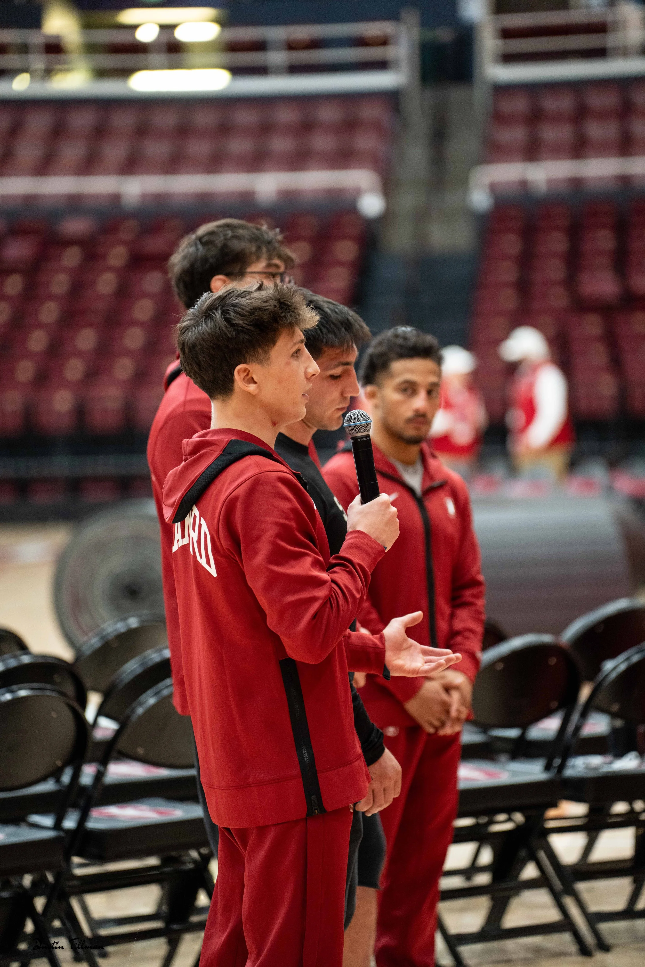 Four young men in red athletic jackets standing in a row inside a gymnasium or sports arena. The man in the foreground is speaking into a microphone, gesturing with his hand, while the others listen attentively. Empty chairs are visible behind them, and the background features a large spiral weight, possibly on a weightlifting machine or storage rack.