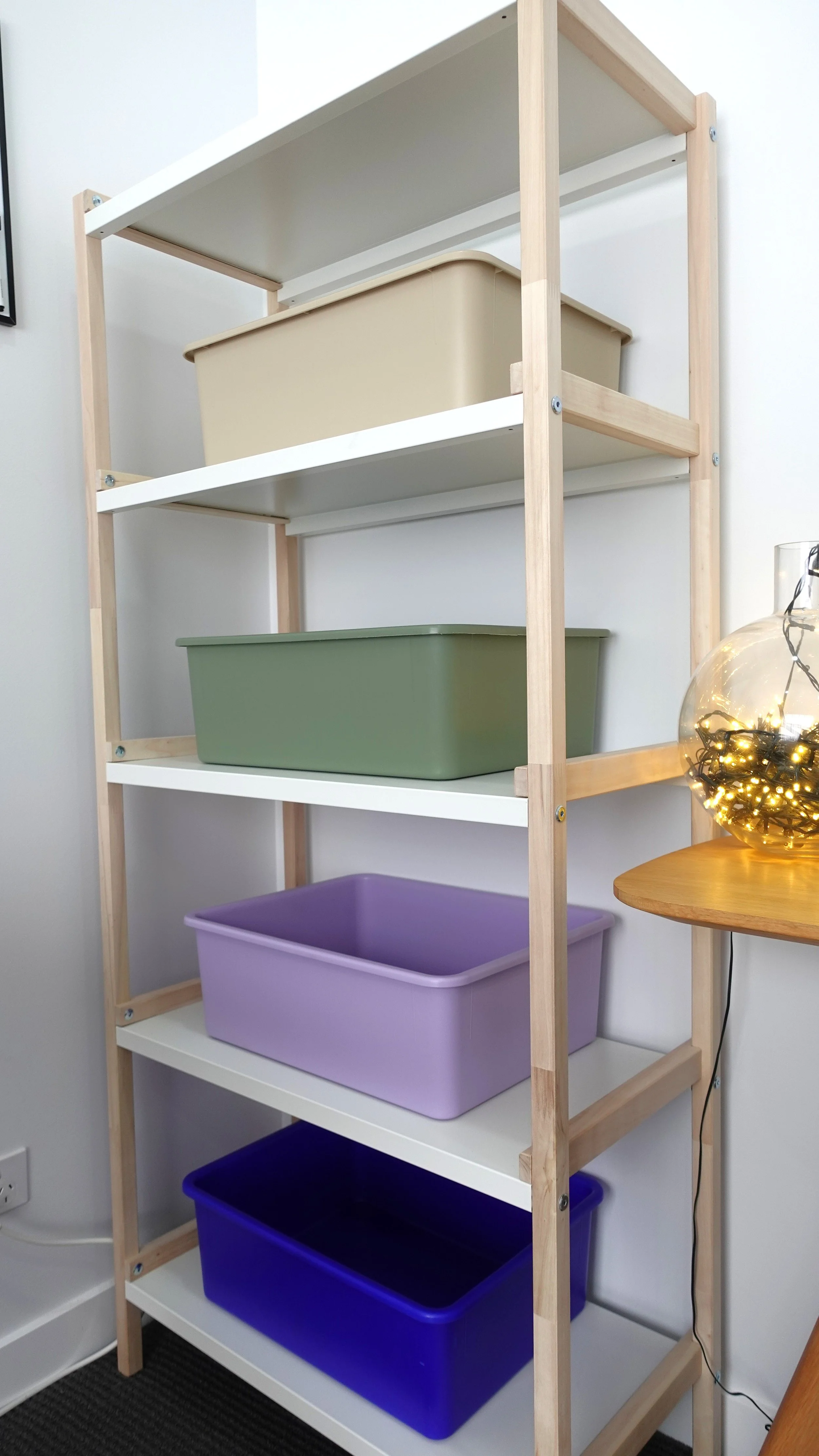 A wooden and white shelving unit with five empty colorful storage bins, against a white wall with a partial view of a lamp with string lights on a small table nearby.