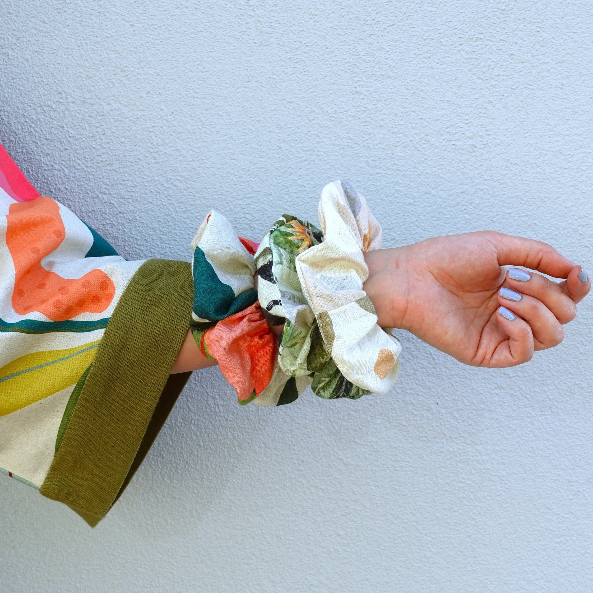 Person wearing a colorful, patterned kimono styled robe with three fabric scrunchie on their wrist, against a plain, light-colored wall.