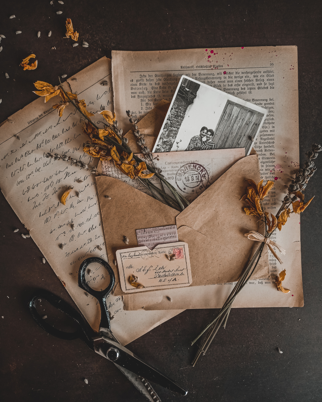Vintage letters, photographs, dried flowers, scissors, and a stamp on a dark surface, creating an old-fashioned, nostalgic scene.