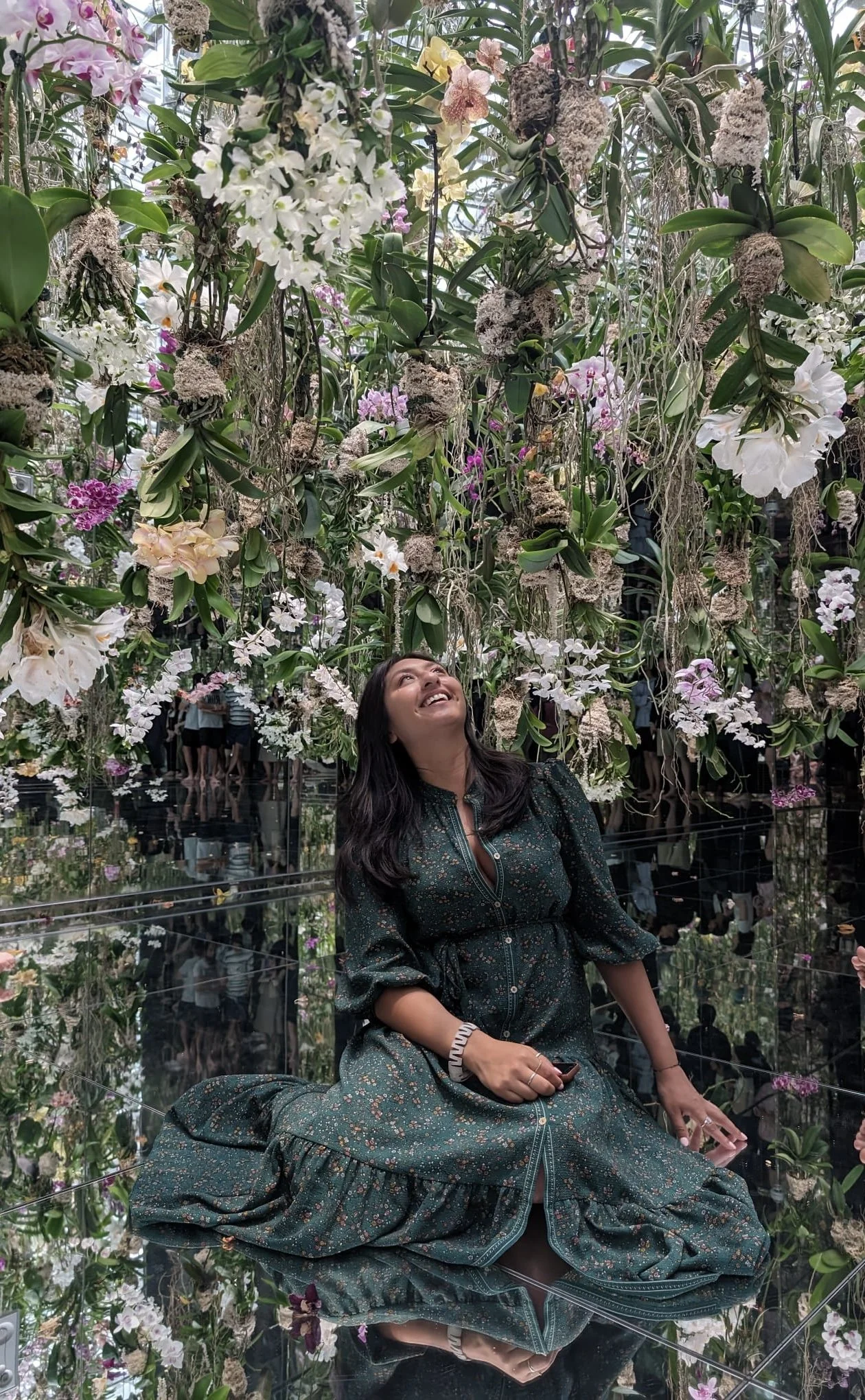 Woman in a green dress sitting on a reflective glass floor, looking up and smiling surrounded by hanging flowers and plants in an indoor botanical garden or orchid exhibit.