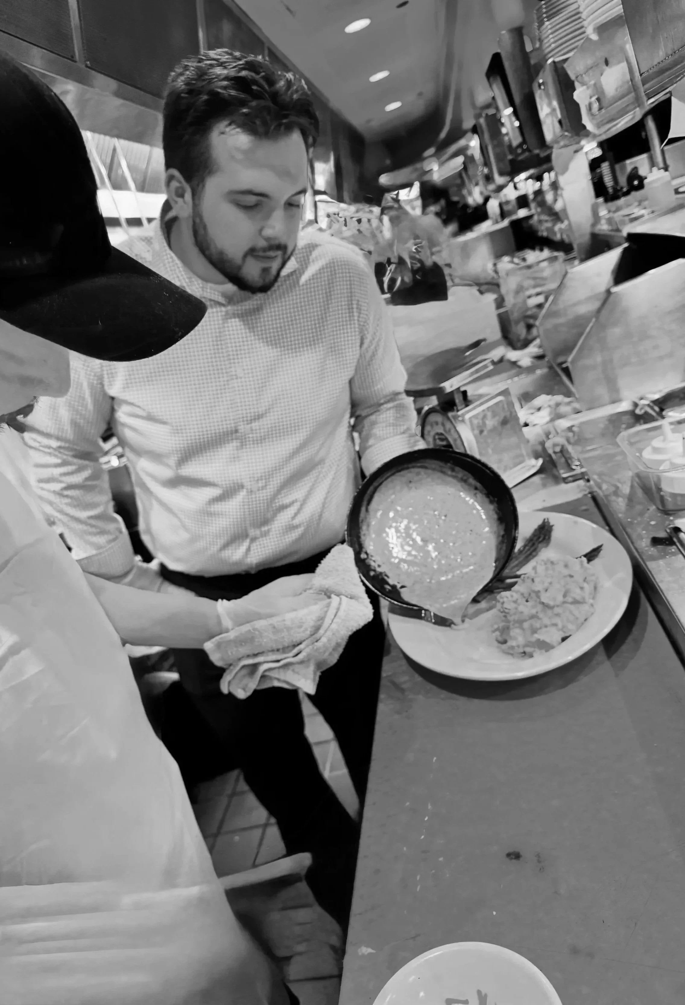 A man in a checkered shirt is pouring gravy onto a plate of mashed potatoes and other food items at a restaurant or diner.