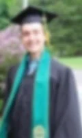 A graduate in a cap and gown with a green sash, standing outdoors on a grass field.