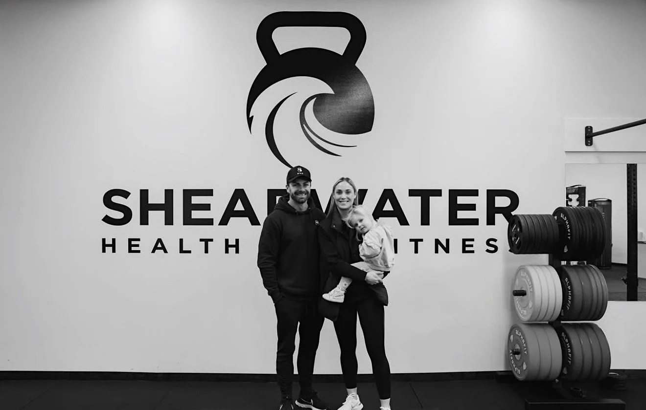 A family of three standing inside a gym with a large 'Shea Water Health & Fitness' logo on the wall behind them. The father, mother, and child are smiling. The gym weights are visible on the right side of the image.