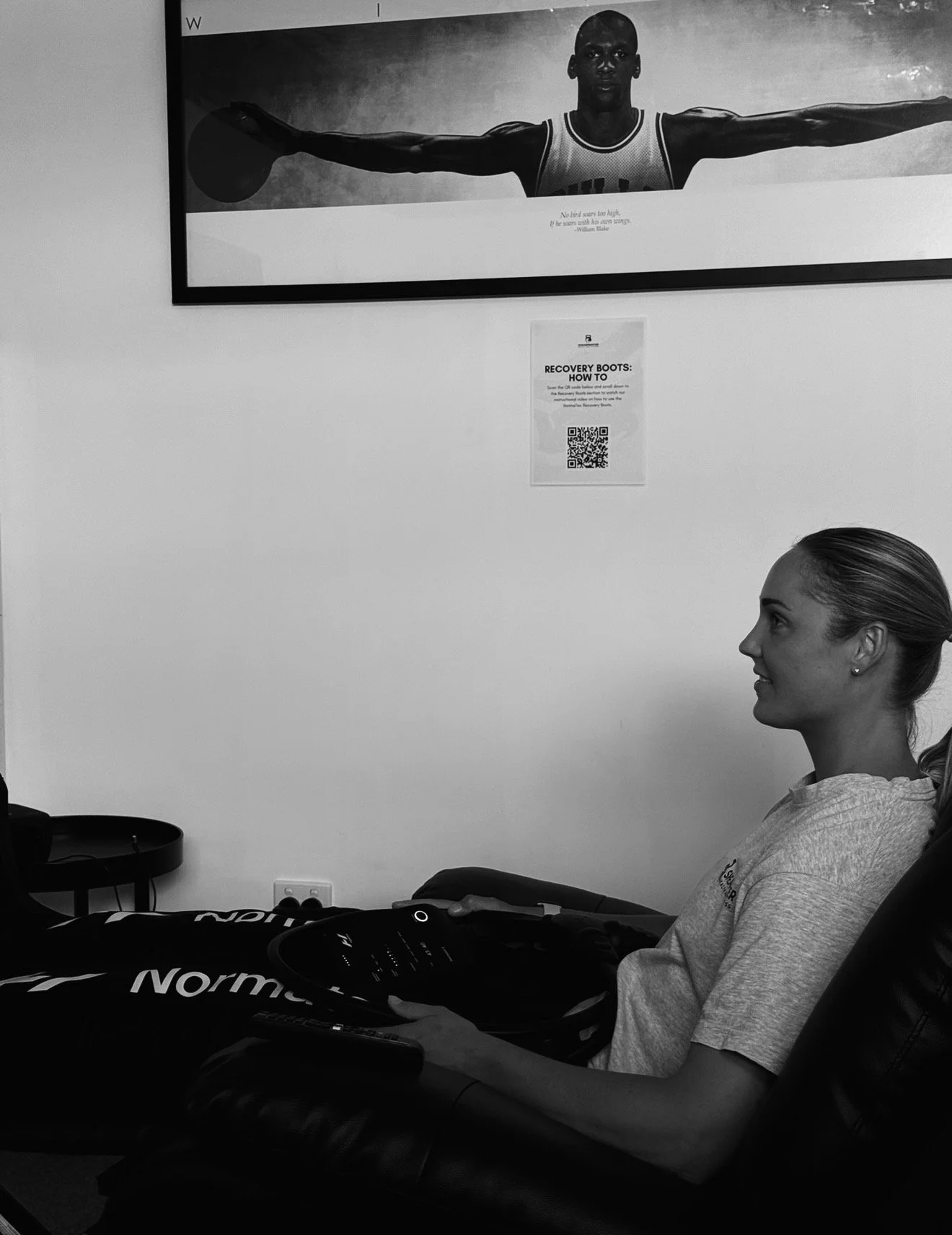 A woman is sitting in a chair at a medical facility, receiving a treatment, with a remote control in her hand. Above her on the wall is a large black-and-white poster of a male athlete holding a basketball with arms outstretched. A smaller sign with a QR code is also on the wall.