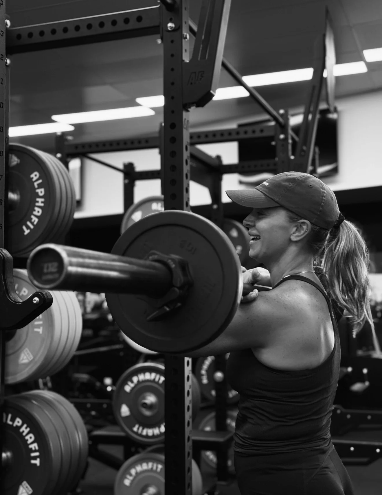 A woman lifting a barbell in a gym, smiling, wearing a cap and workout clothes, with weight plates in the background.
