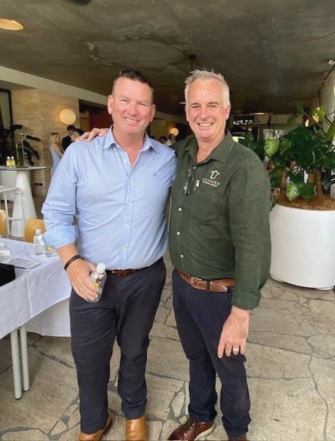 Smiling buyers agent Marty Ryan wearing a green shirt with 'Lilyburn Buyers Agency' embossed, glasses hanging from his collar, standing indoors with posters and photos in the background.