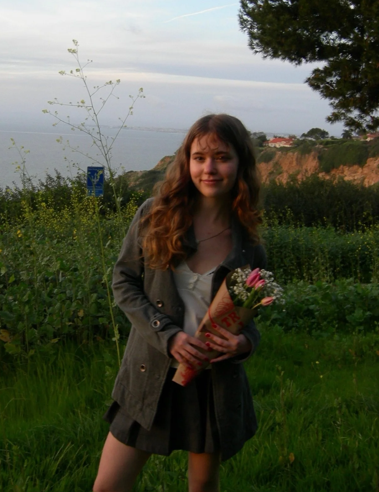Photograph of Lucy Caliri, a young woman with long, wavy brown hair. She's holding a bouqet of pink roses and white baby's breath, standing outdoors with scenic coastal cliffs in the background.