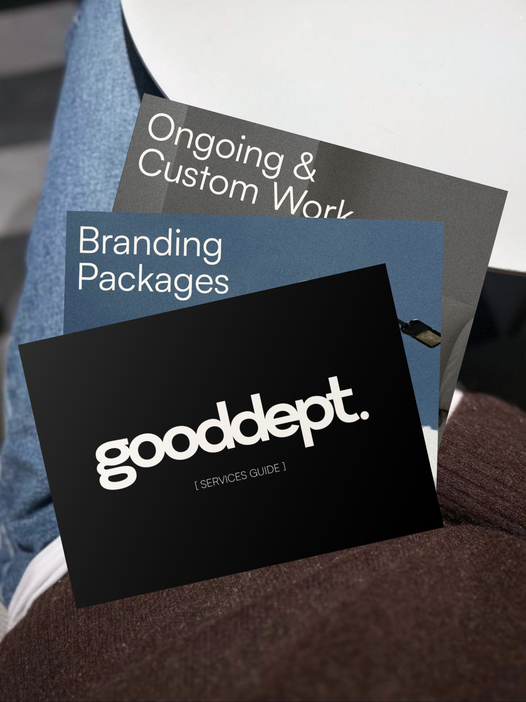 A person holding a black booklet with white text that says 'gooddept. SERVICES GUIDE' and additional booklets behind it with titles 'Ongoing & Custom Work' and 'Branding Packages'.