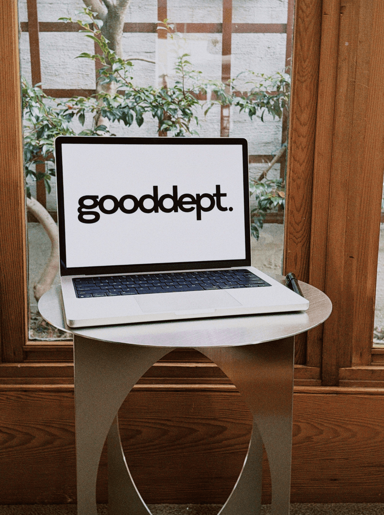Laptop on a small table with the word 'gooddepot.' on the screen, set in a cozy space by a window with plants outside.