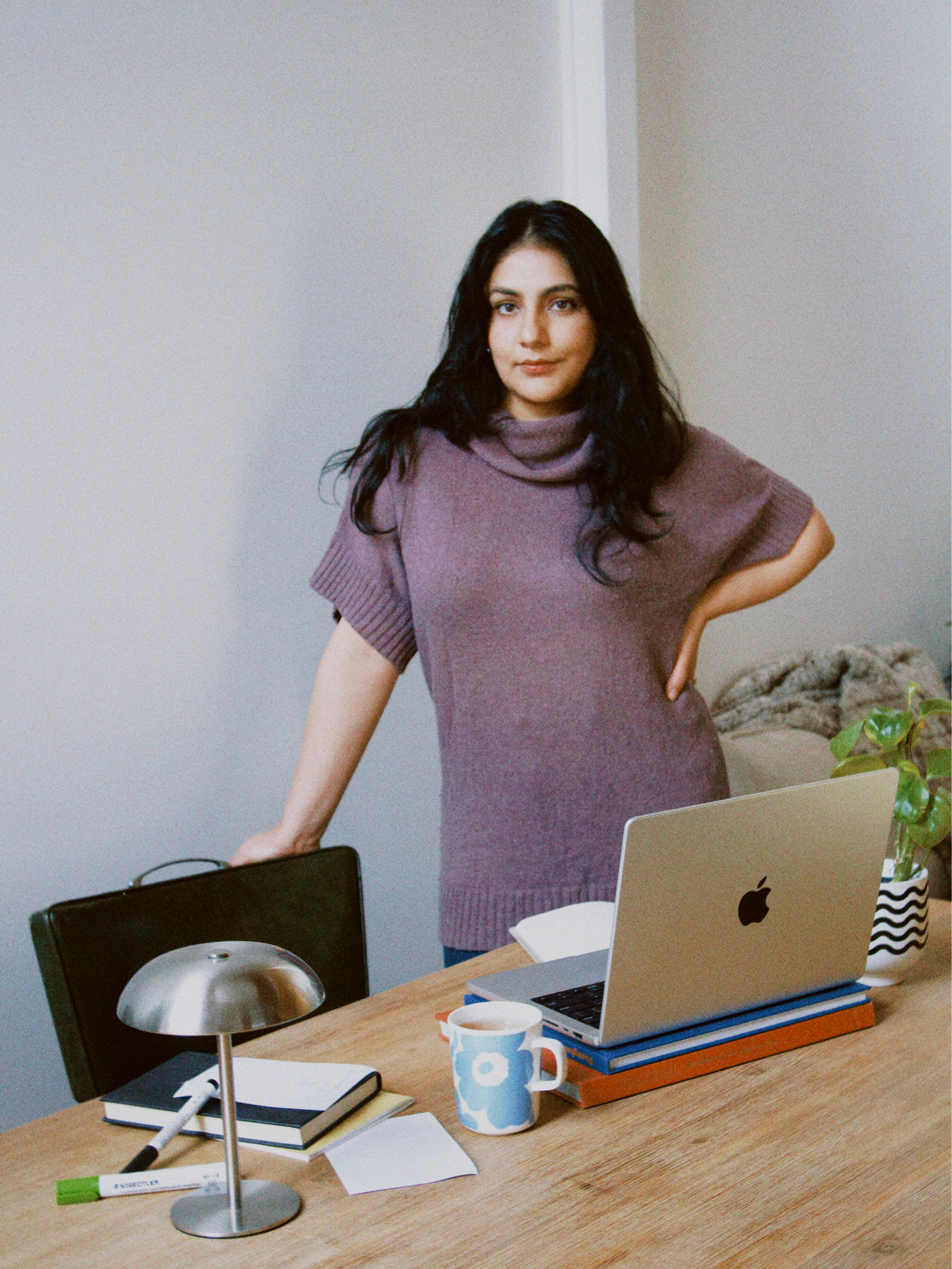 woman Nikita Lamba with dark hair standing in an office with a wooden table, a laptop, a mug, and office supplies.