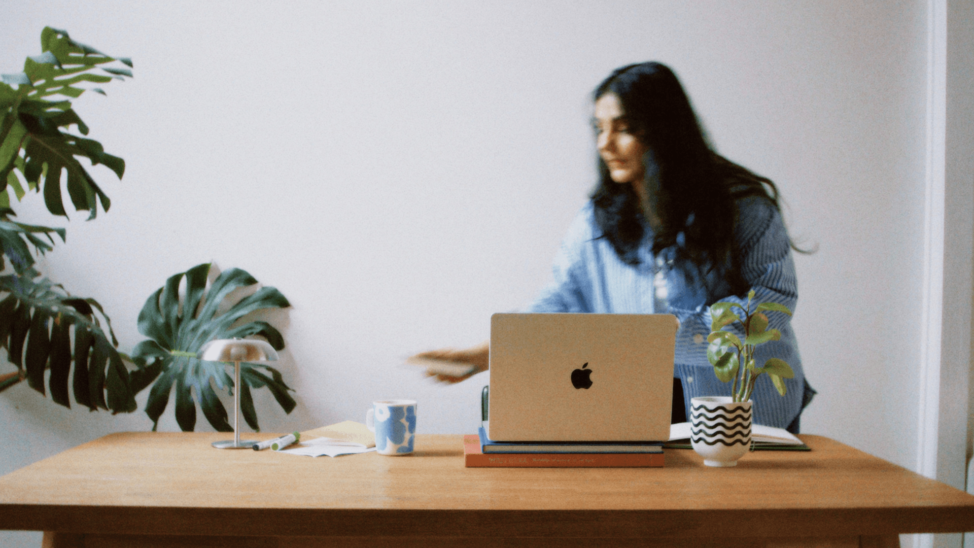 A woman with long dark hair working at a wooden desk with a silver laptop, books, a mug, and decorative plants, in a bright room with white walls.