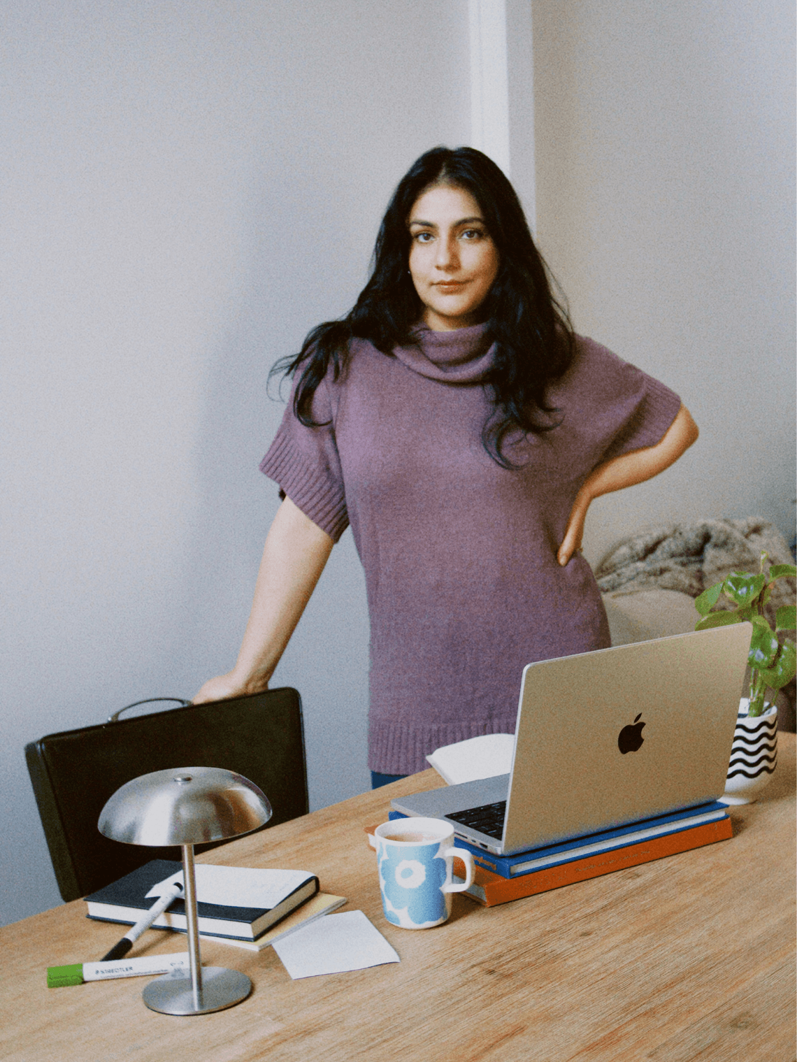 woman Nikita Lamba with dark hair standing in an office with a wooden table, a laptop, a mug, and office supplies.