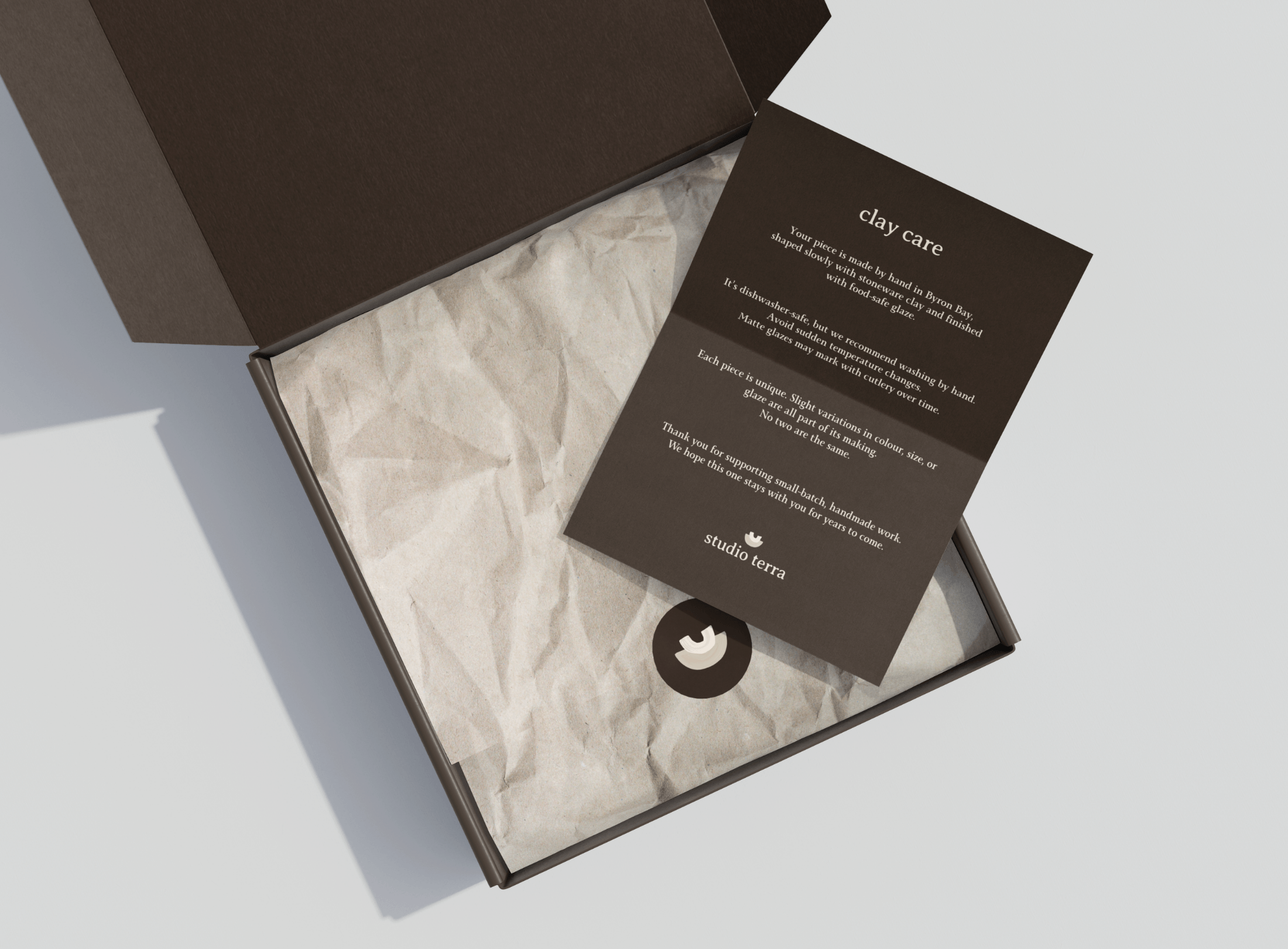 Open brown box lined with crumpled paper, with a black card lying inside explaining clay care instructions from Studio Terra.