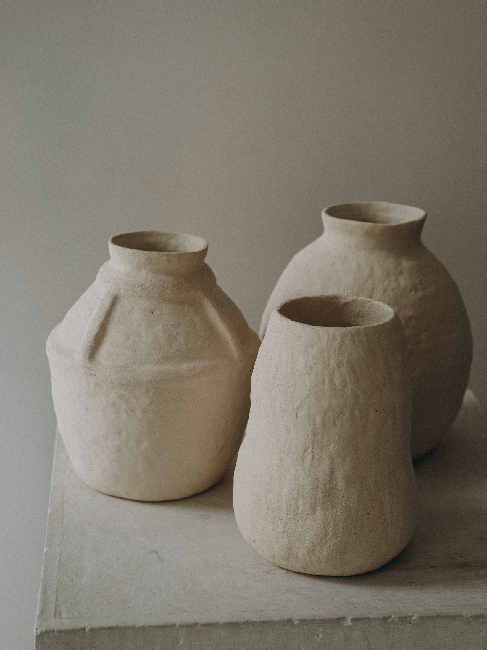 Three handmade ceramic vases with natural, unglazed, textured surfaces placed on a light-colored rectangular platform with a plain background.
