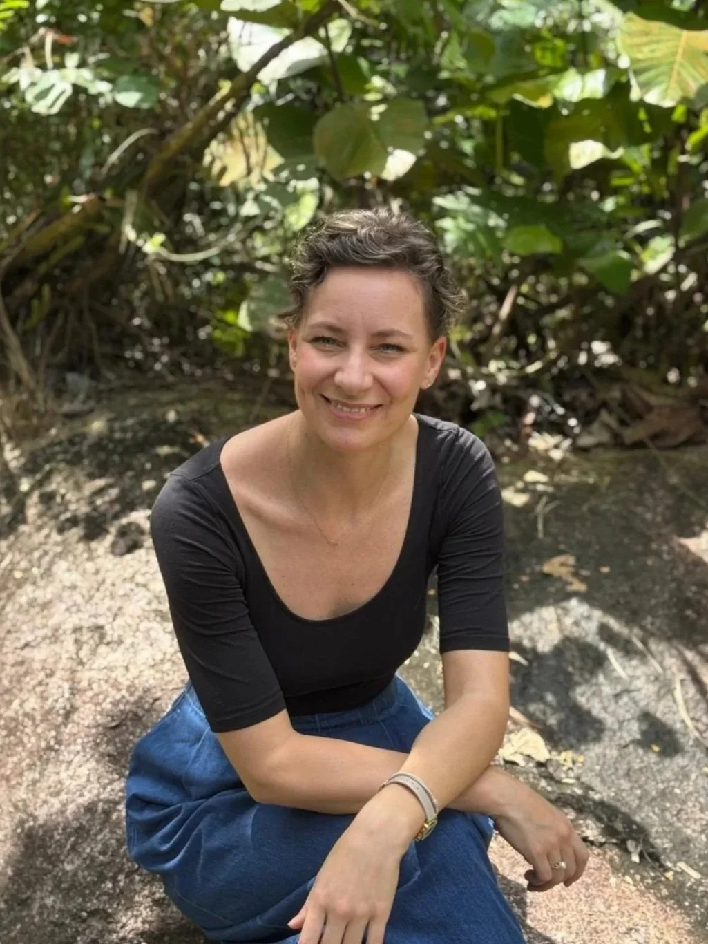 A woman with short dark hair sitting outdoors in a green, leafy environment, smiling at the camera.