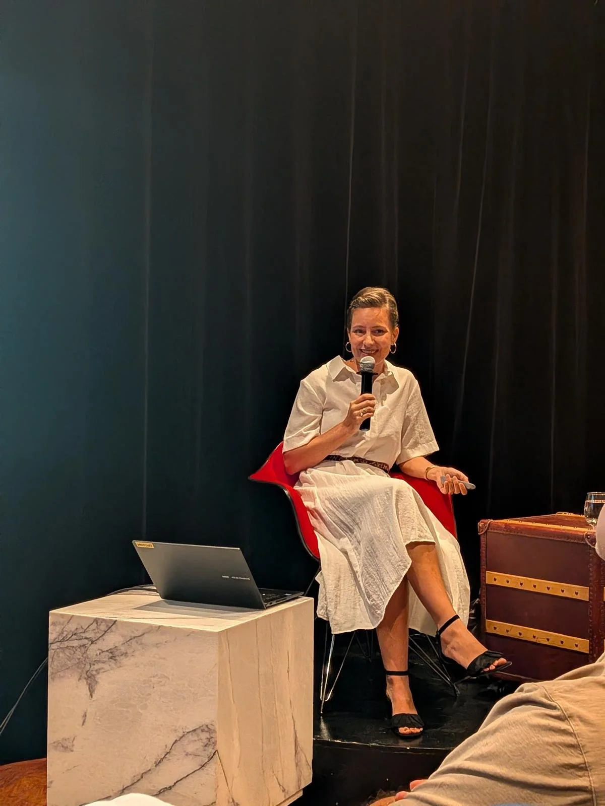 Woman speaking into a microphone while seated on a red chair at talk, with a laptop on a marble side table and a dark background.