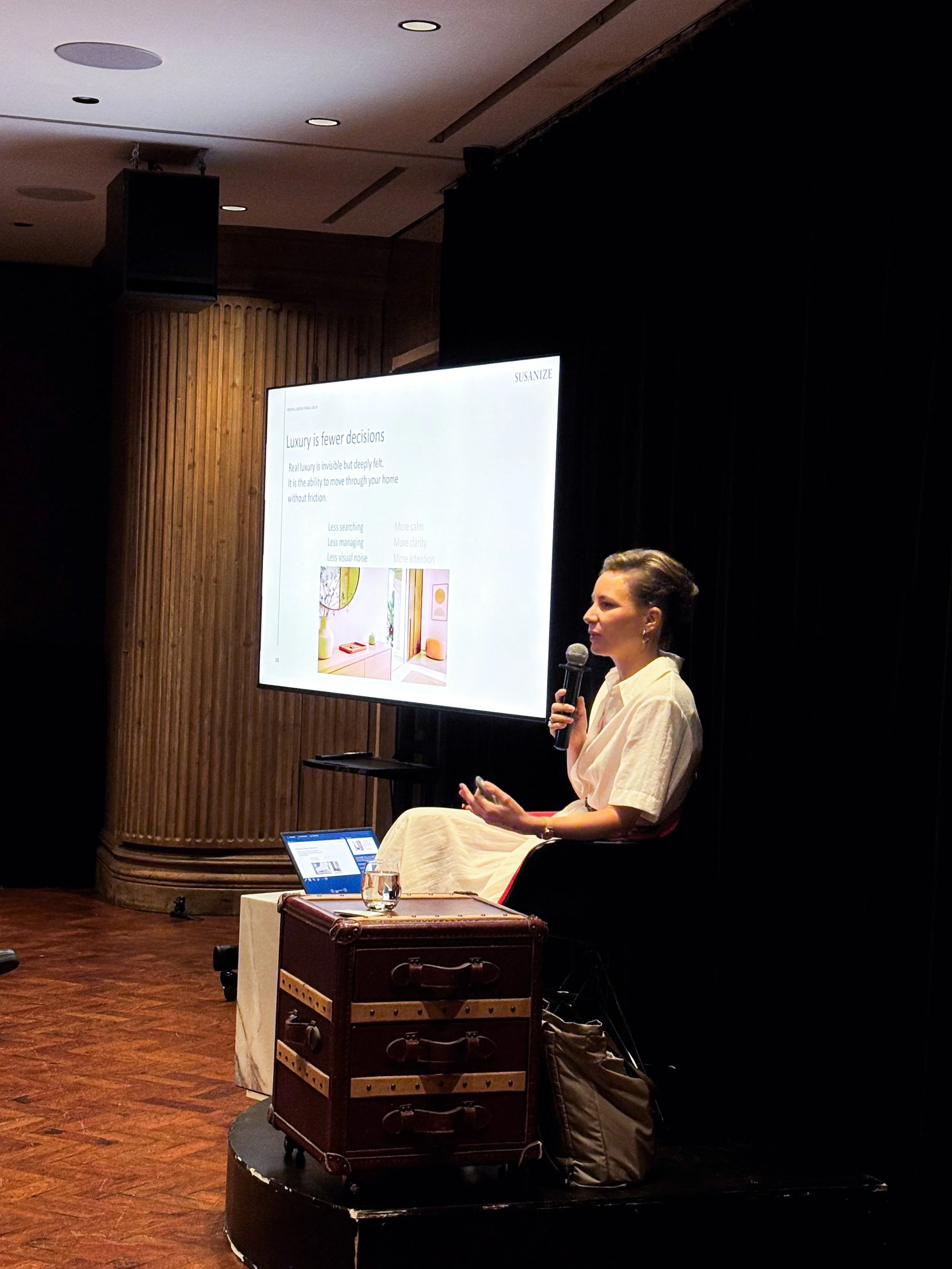 A woman in a light-colored dress seated on a stage, speaking into a microphone. Behind her is a large presentation screen displaying text about luxury and home design, with a laptop on a small table nearby.